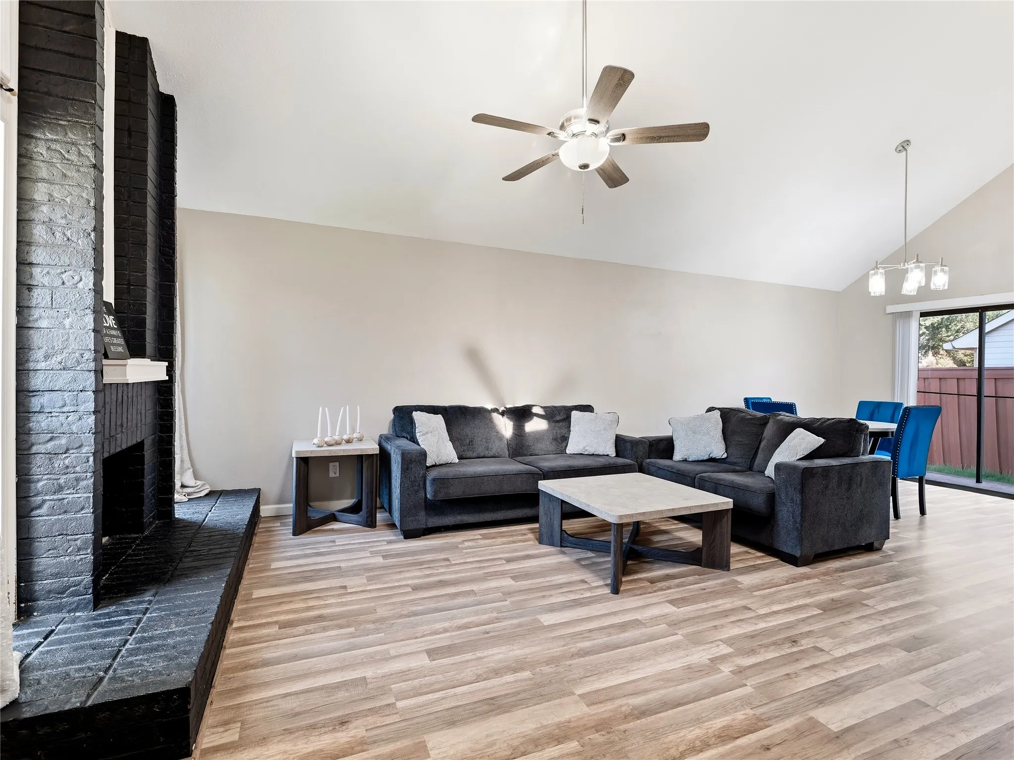 Vaulted ceilings, a dark brick fireplace, wood-finish flooring, a ceiling fan, and a sliding glass door