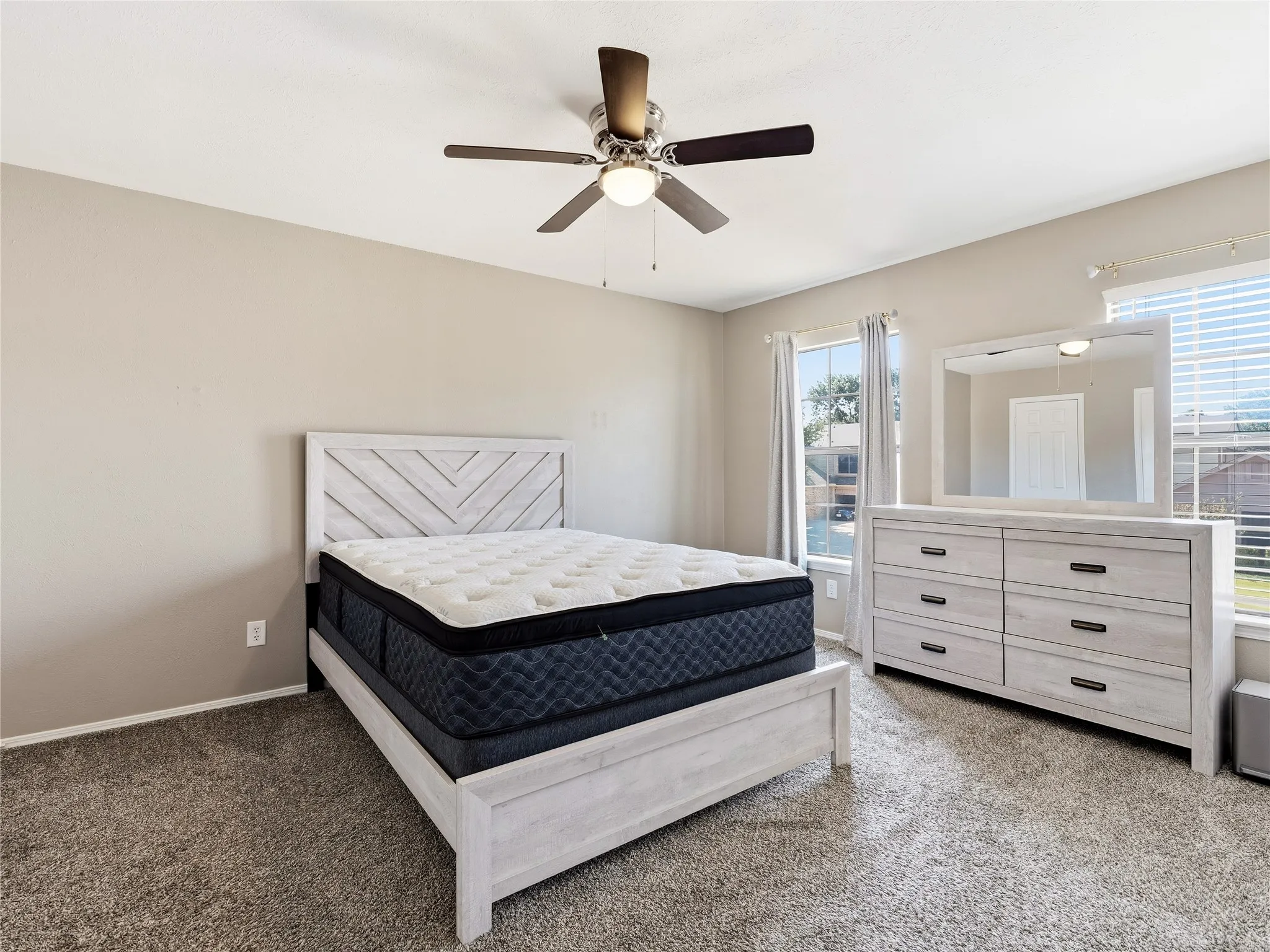 Soft neutral wall tones, textured carpeting, two windows with blinds, and a ceiling fan with integrated lighting