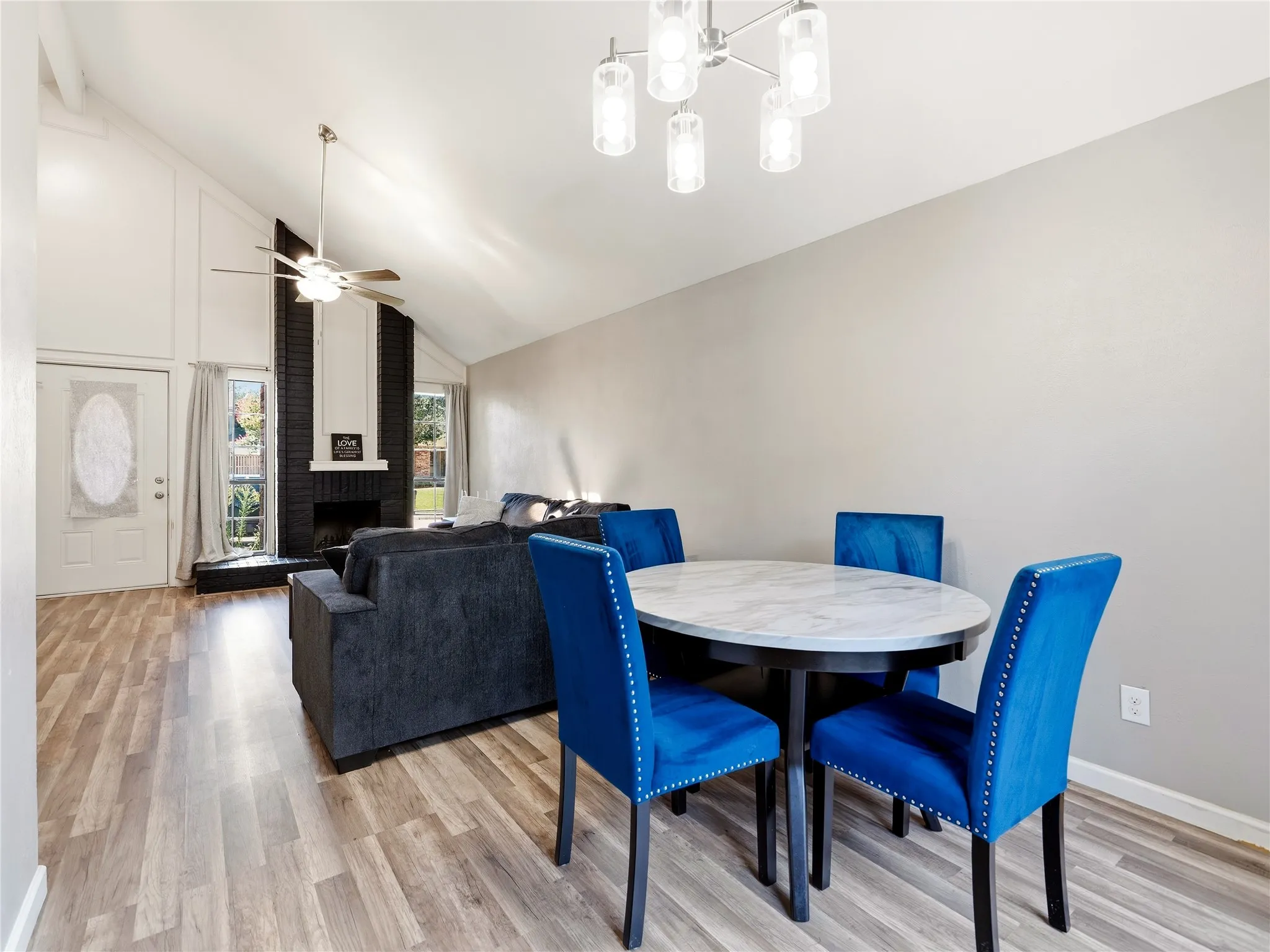 Vaulted ceiling with exposed beam, wood-finish flooring, black brick fireplace with white mantel, white door with oval decorative glass insert, and modern ceiling light fixture