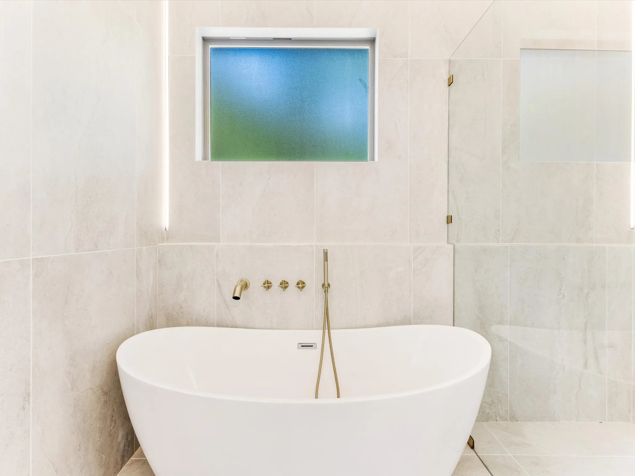 Freestanding soaking tub with a wall-mounted brass faucet, floor-to-ceiling tiled walls, and an integrated glass shower partition