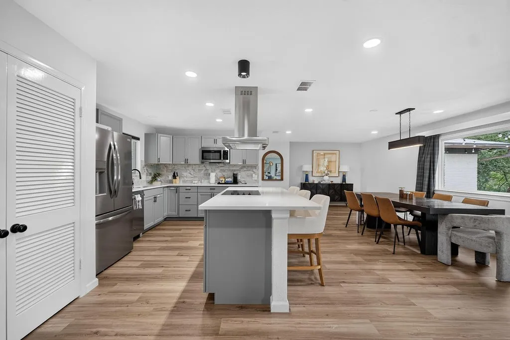 Open-concept living area with wood-finish flooring, a kitchen featuring light gray cabinetry and stainless steel appliances, a center island with a white countertop, and recessed lighting