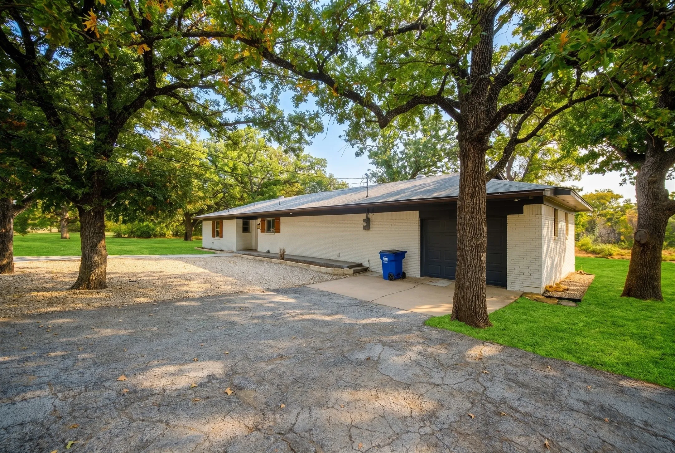 White painted brick exterior with dark gray garage doors, mature trees, a gravel driveway, and a concrete pad