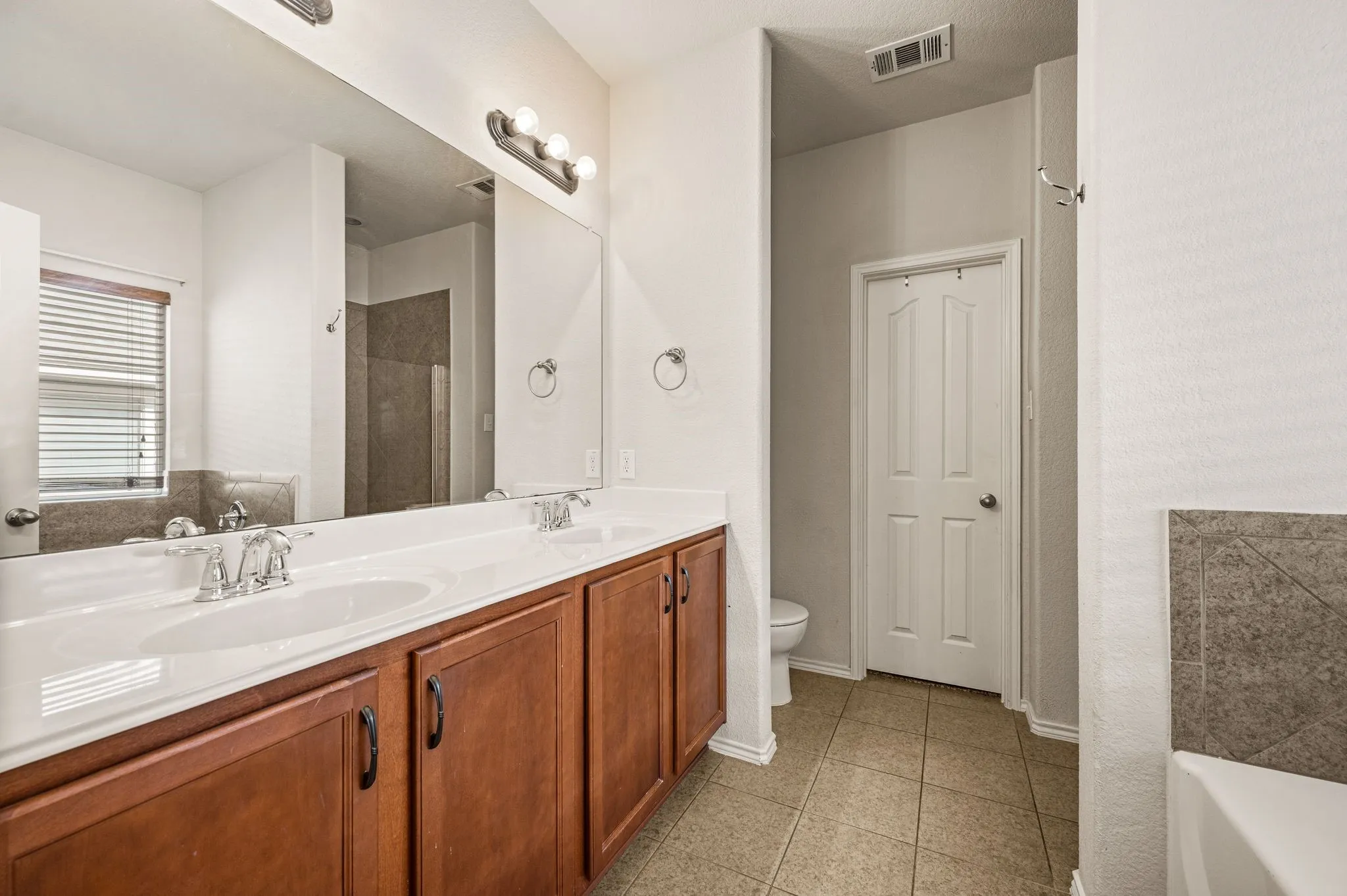 Dual vanity with integrated sinks, wood-finish cabinetry, a large wall-mounted mirror, and tile flooring