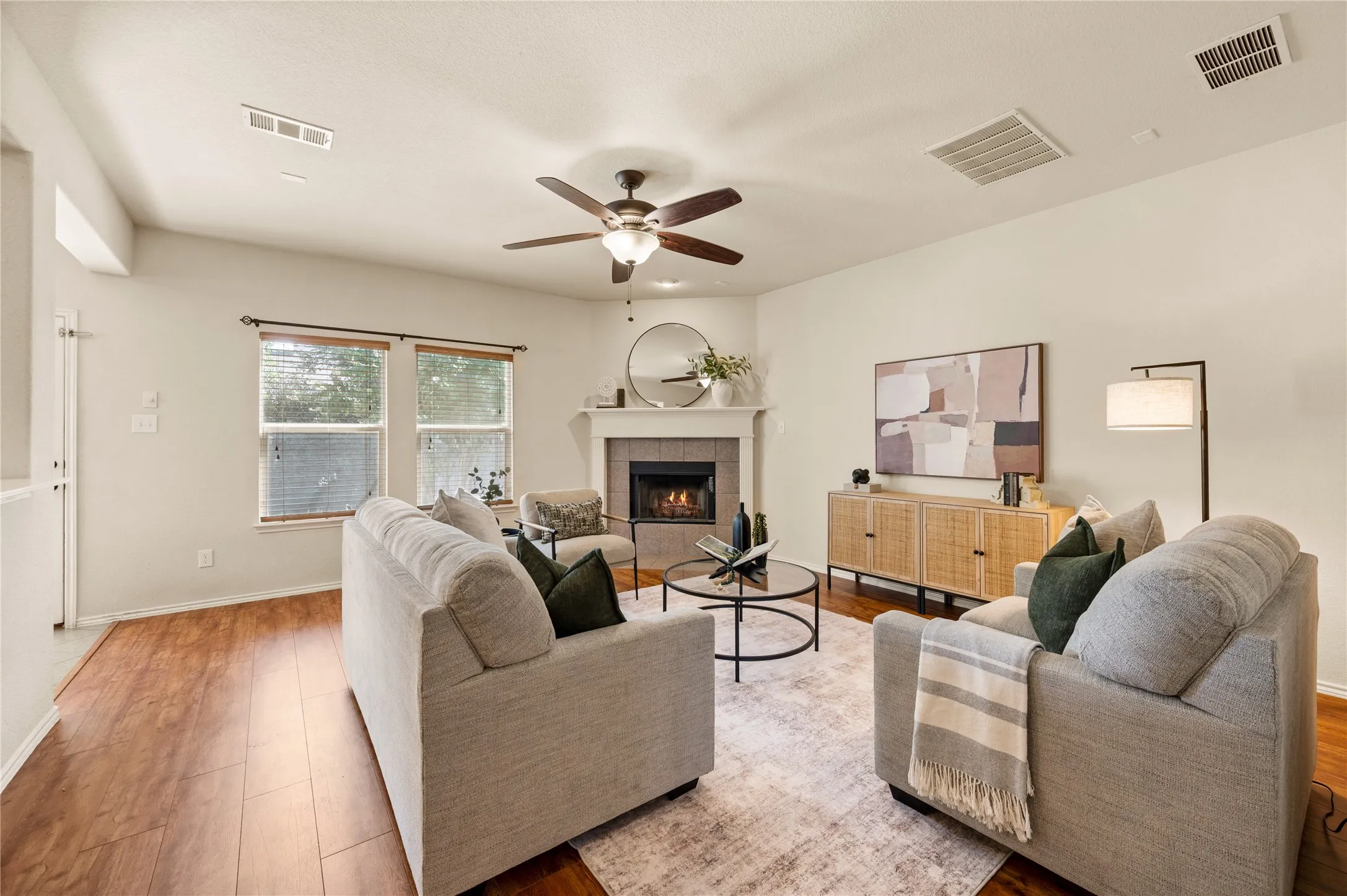 Living area with wood-finish flooring, two large windows, a corner fireplace with a mantel, and an overhead ceiling fan