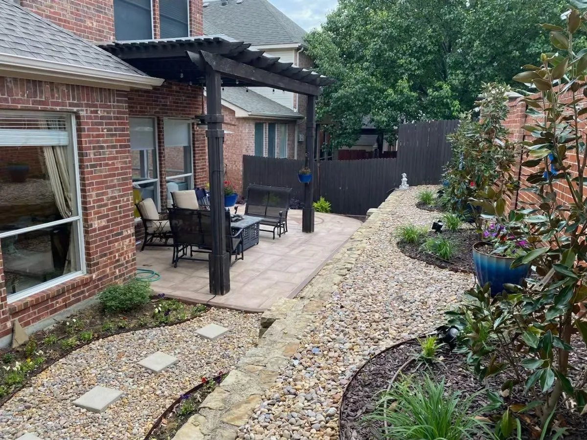 Brick exterior with multiple windows, paver patio, dark wood pergola, gravel pathways with stone retaining walls, and planted garden beds