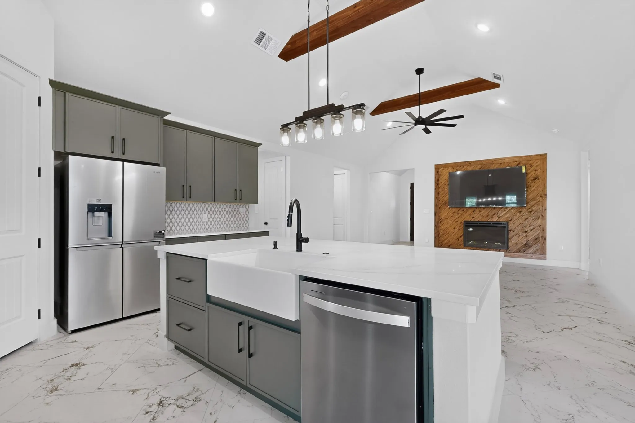 Vaulted ceiling with exposed wood beams, white marble-look flooring, central island with white farmhouse sink, stainless steel appliances, and a wood-paneled accent wall with an integrated fireplace