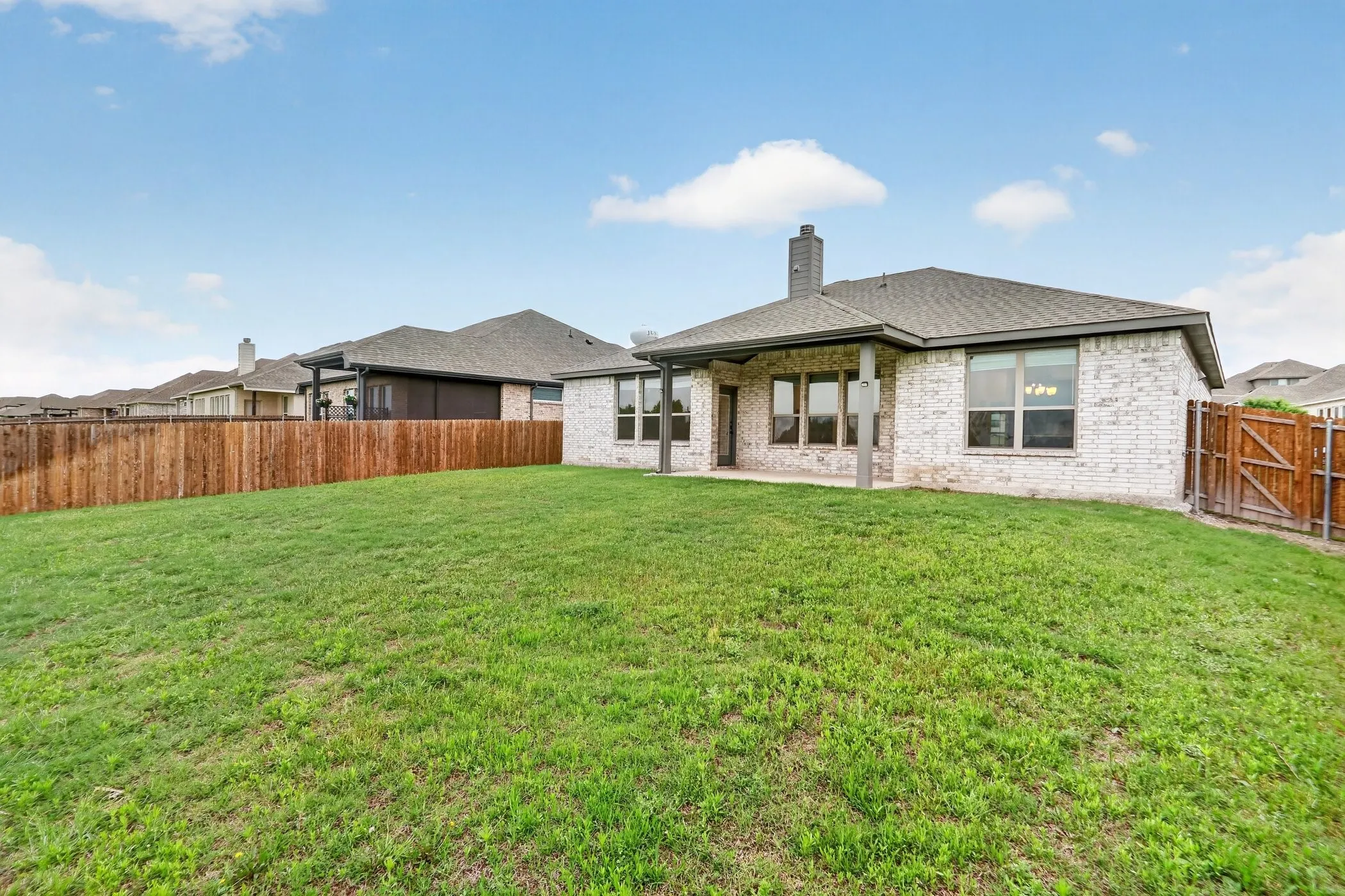 Expansive green lawn, white brick exterior, covered patio, dark shingle roof, and a wood privacy fence