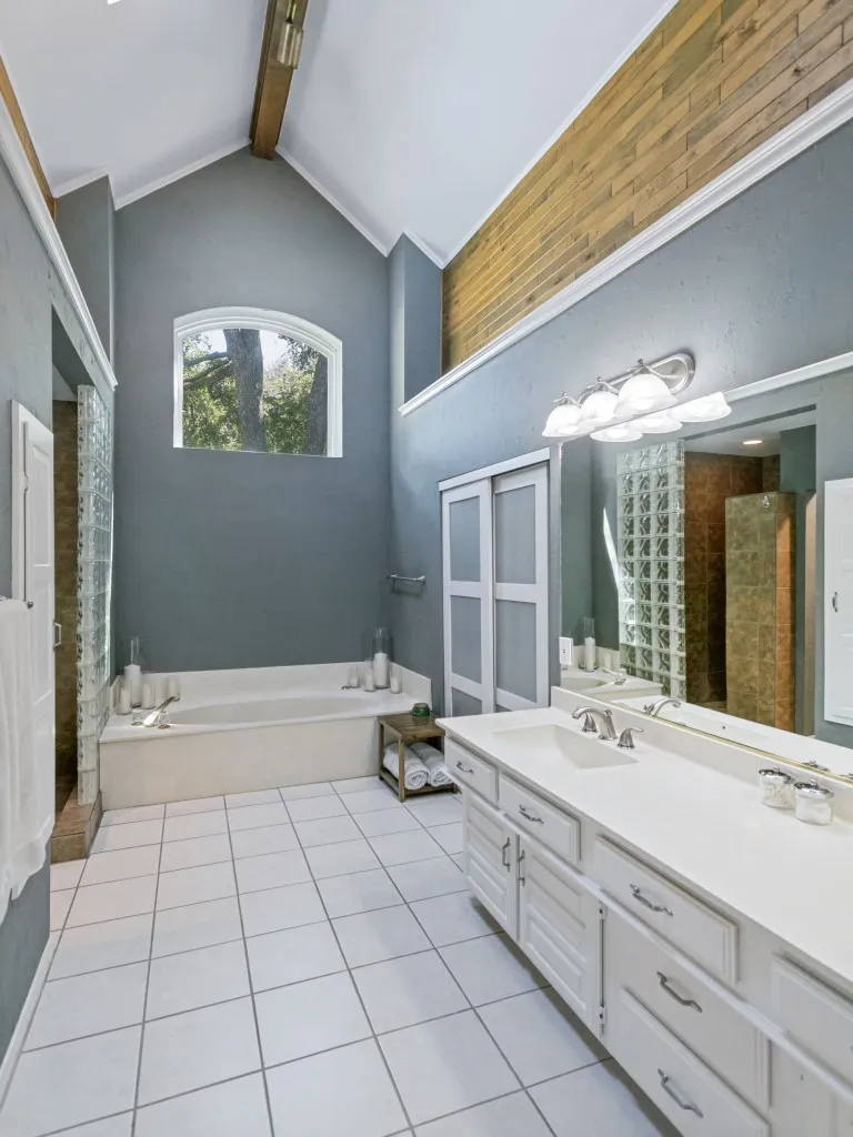 Dramatic primary bathroom features vaulted ceiling with an exposed wood beam, arched window, built-in soaking tub, dual vanity, separate shower and two closets.