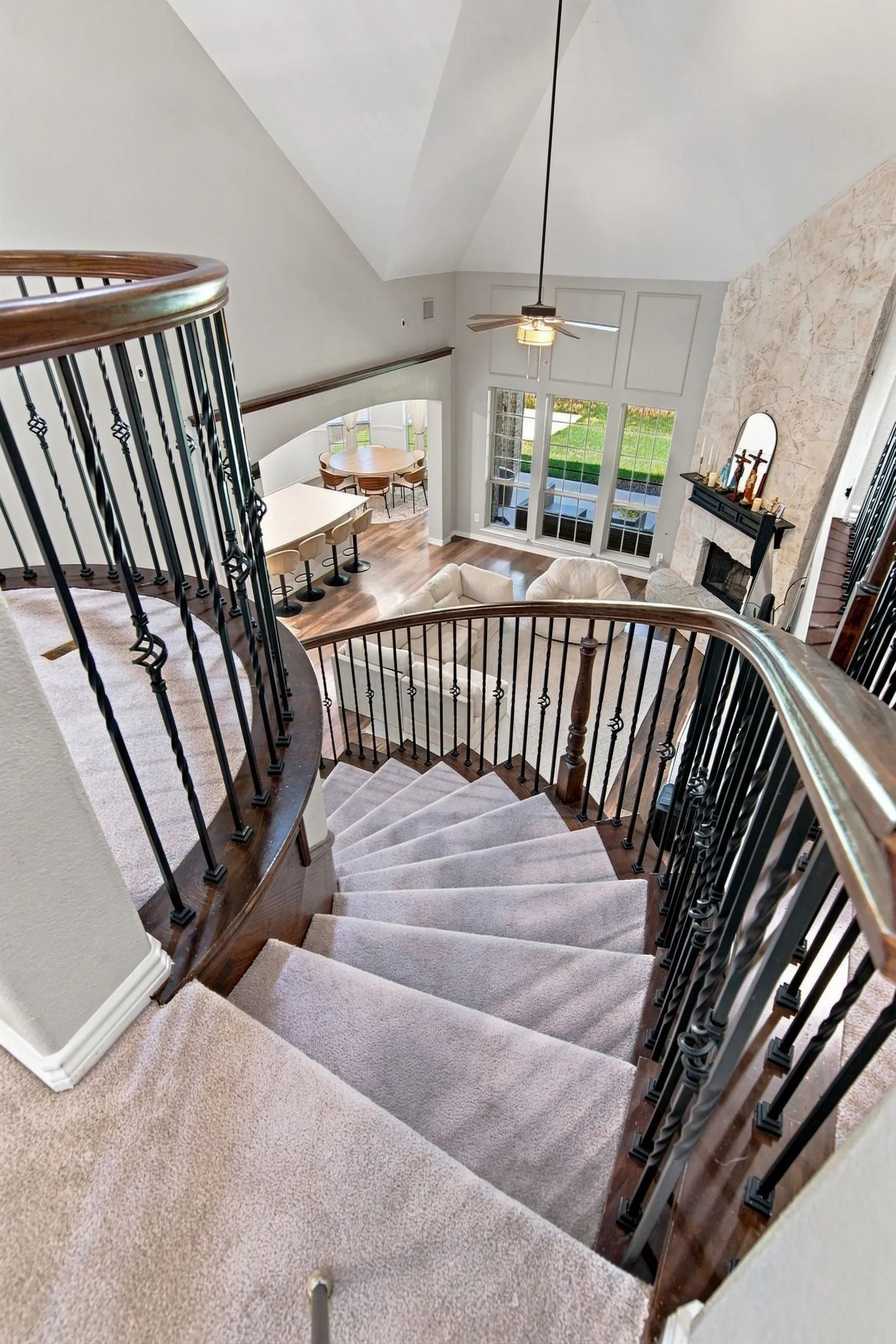 Curved staircase with wrought iron balusters and wood handrails, vaulted ceiling, large windows, and a stone-clad fireplace