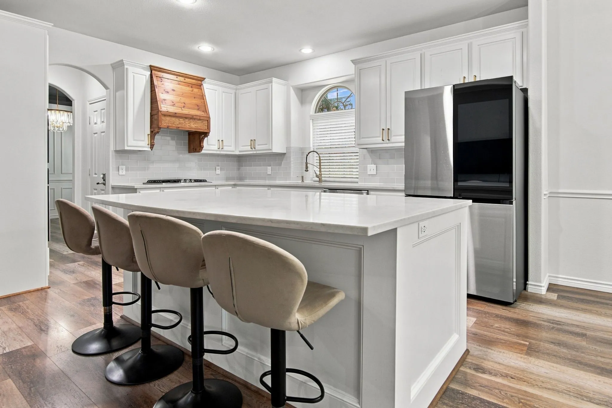 Kitchen featuring a large island with white paneling, white cabinetry with gold-tone hardware, wood-finish flooring, a wood range hood, and a stainless steel refrigerator