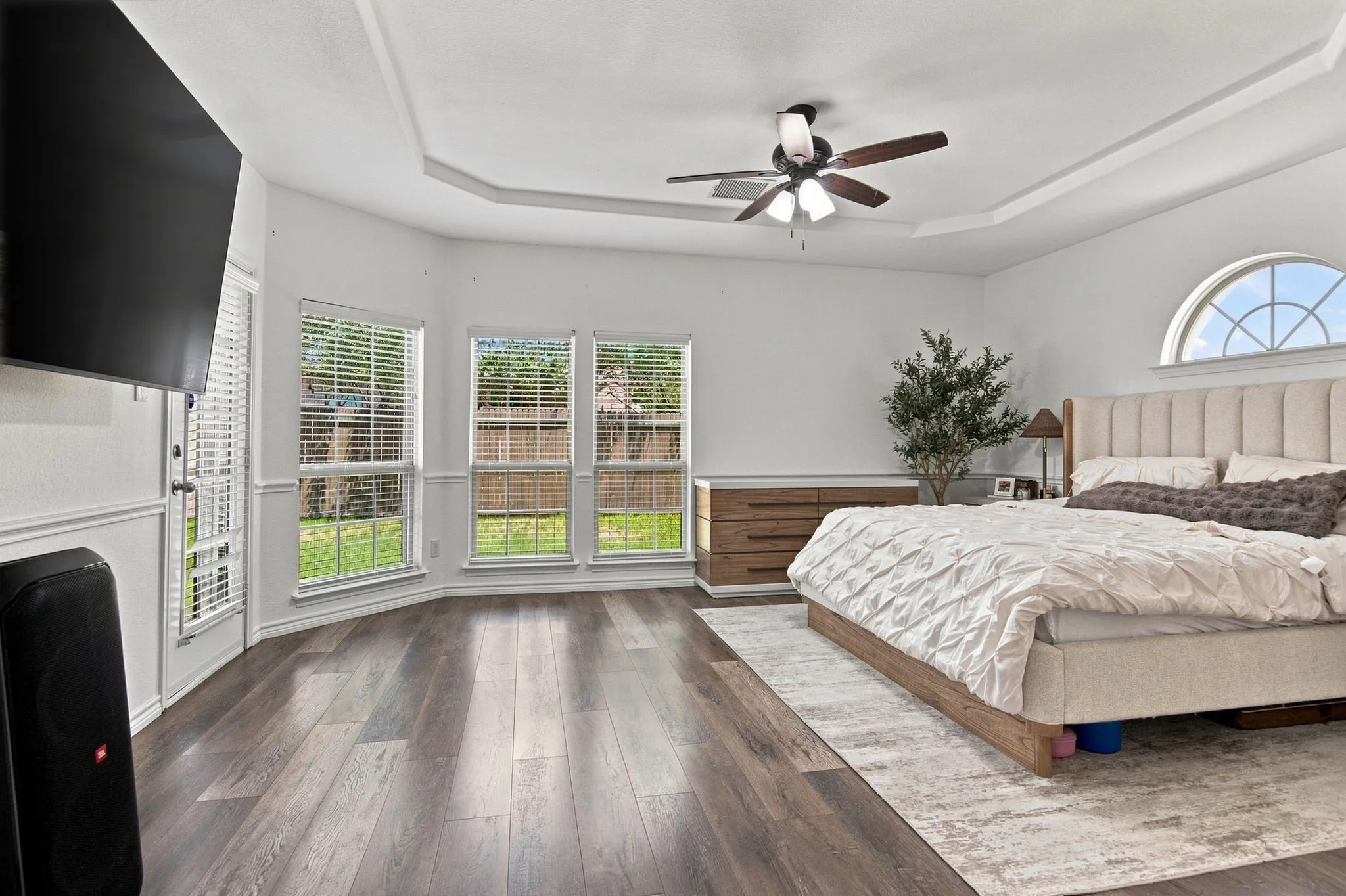 Tray ceiling, ceiling fan, wood-finish flooring, half-round window, and multiple windows with blinds