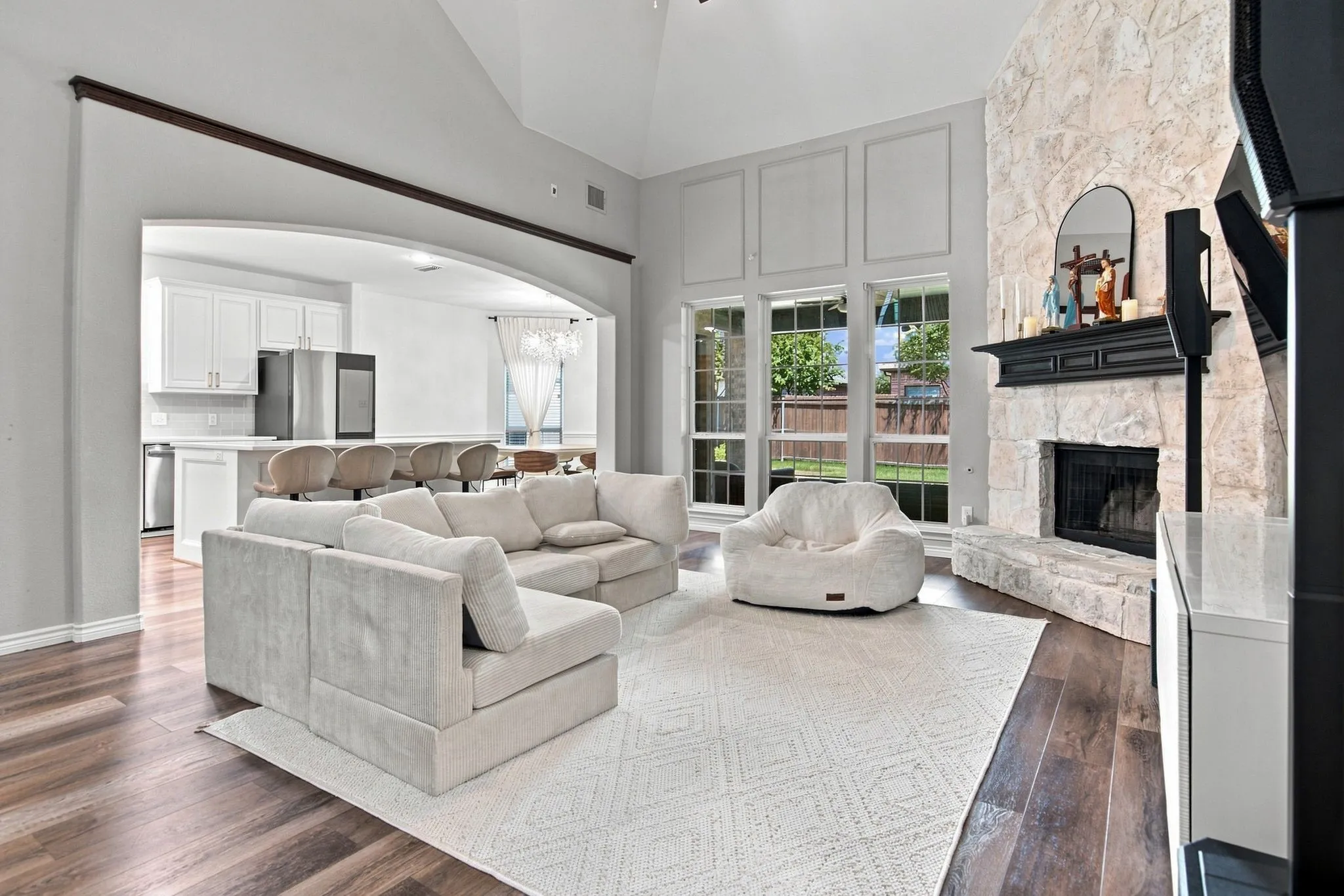 Vaulted ceiling, stone fireplace with dark wood mantel, large windows, wood-finish flooring, and an arched entryway