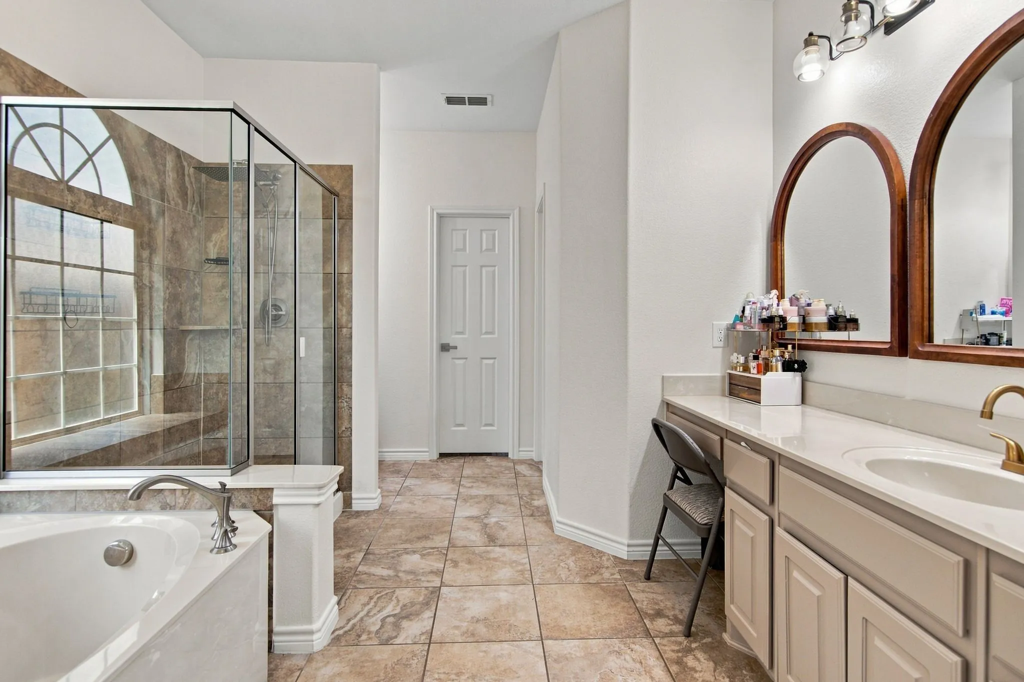 Tiled flooring throughout, soaking tub with deck-mounted faucet, glass-enclosed shower with tiled surround, dual vanity with undermount sinks, and arched mirrors