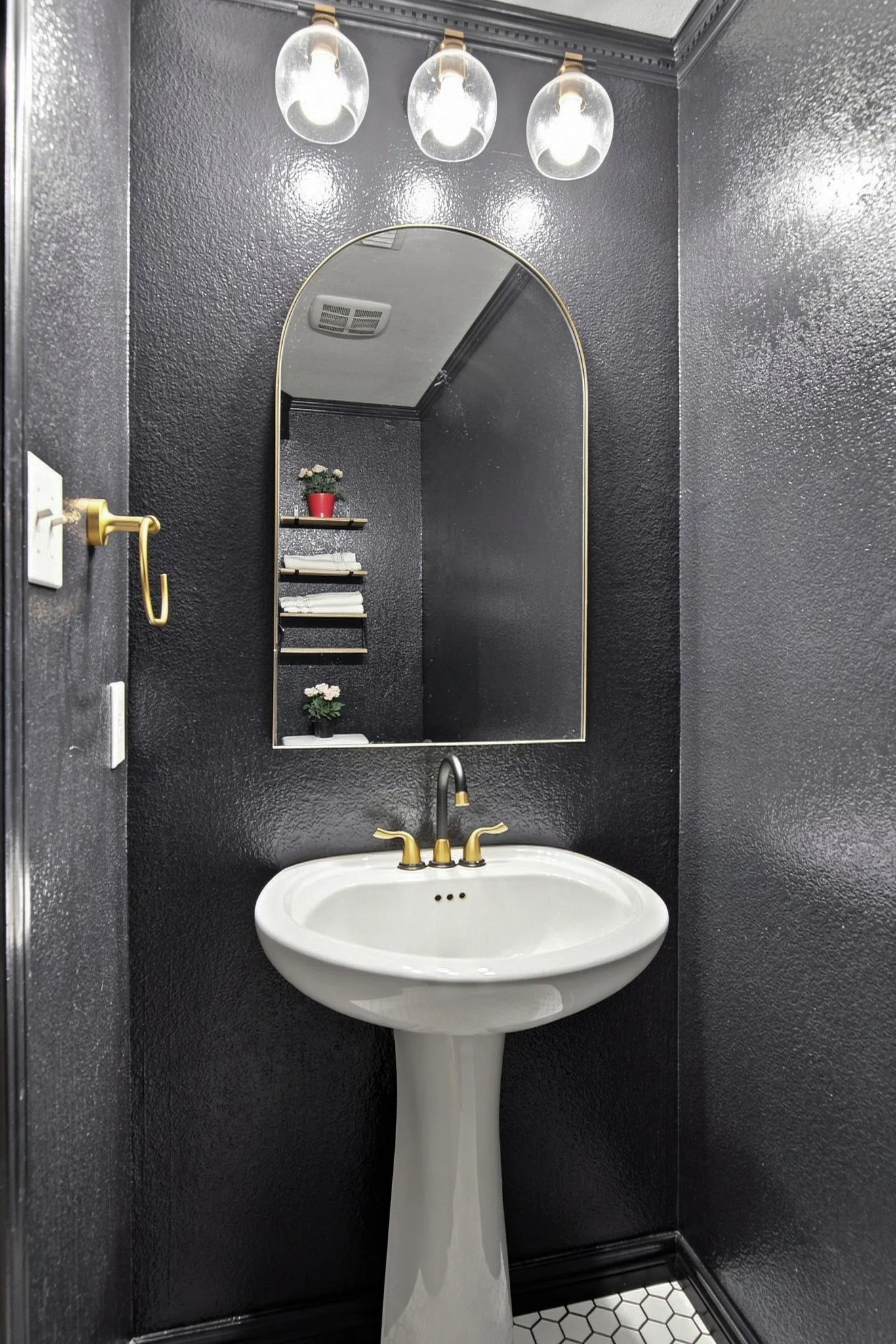 Pedestal sink with brass-finish faucet, arched mirror, globe vanity lighting, and hexagonal tile flooring