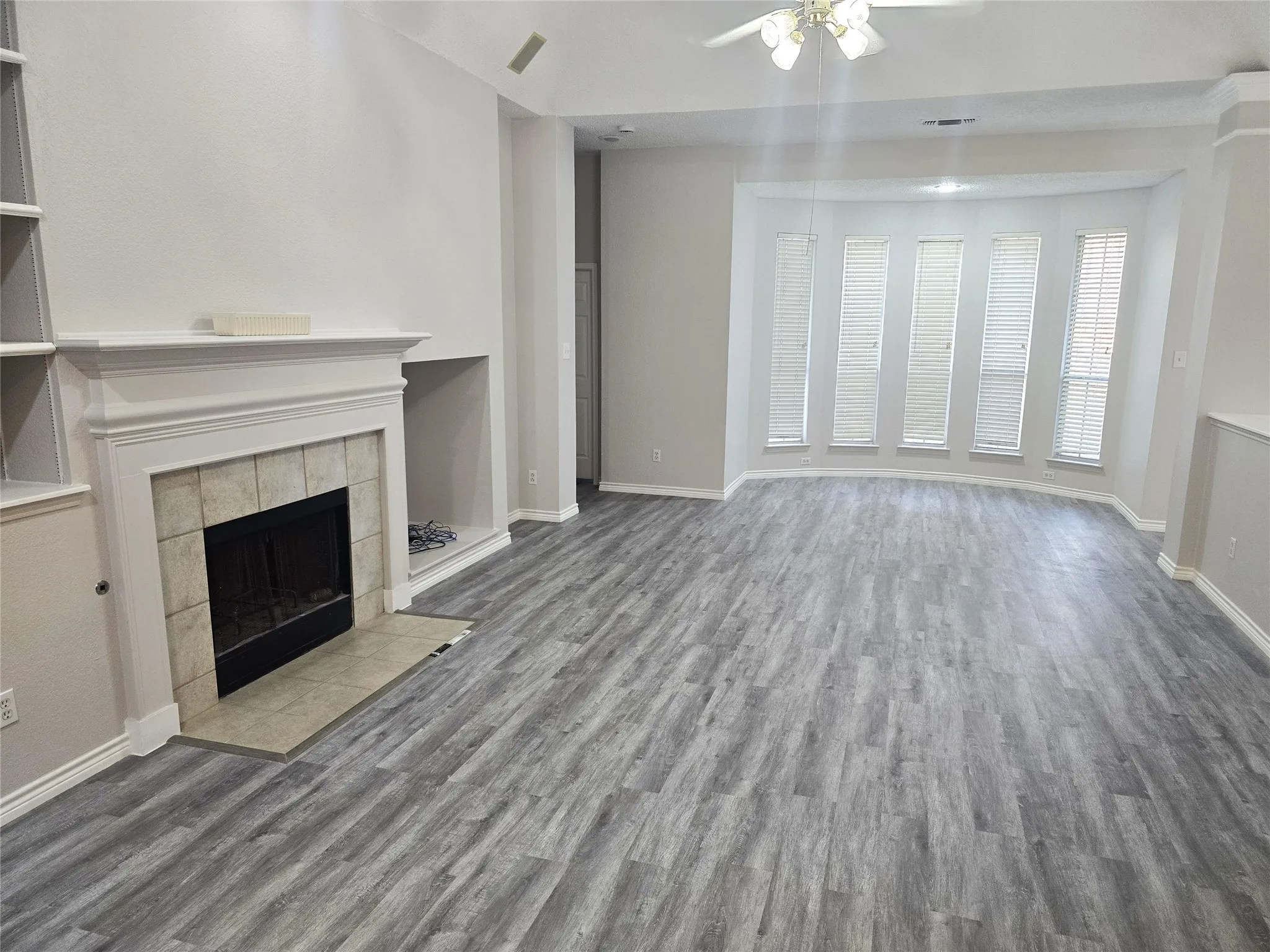 Spacious living area featuring wood-finish flooring, a fireplace with tile surround, a built-in shelving unit, and multiple windows with blinds