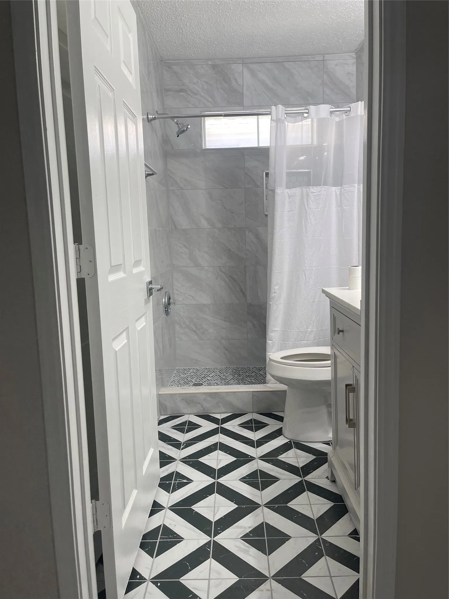 Black and white geometric tile flooring, walk-in shower with light gray tile surround, shower window, white paneled door, and white vanity with cabinet storage