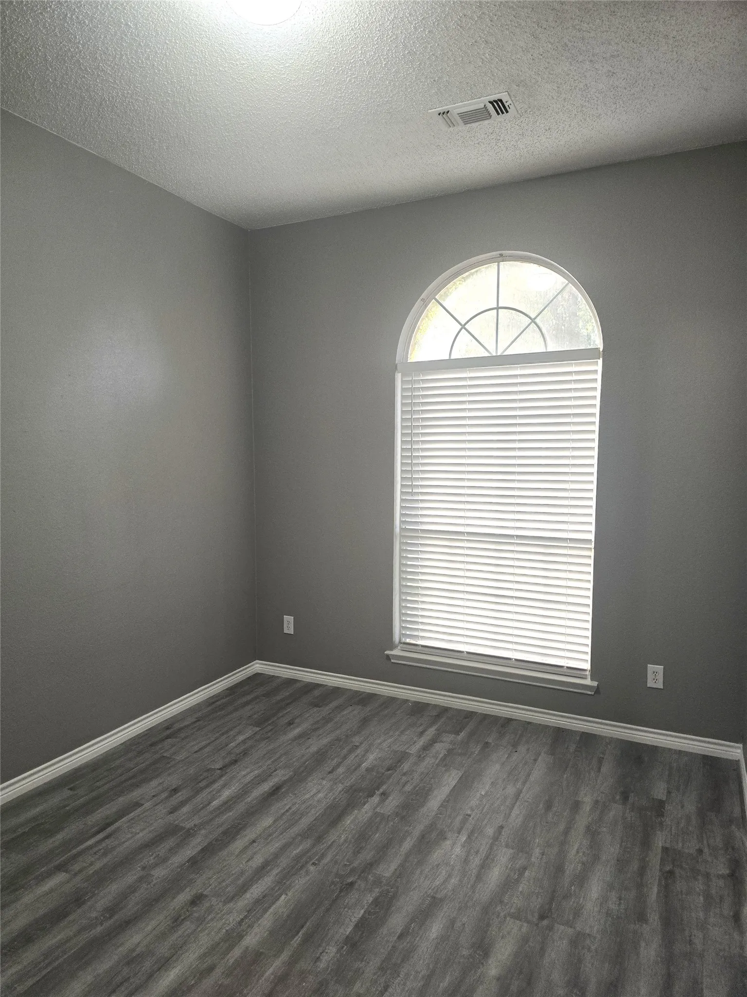 Arched window with blinds, gray wall finish, and wood-finish flooring