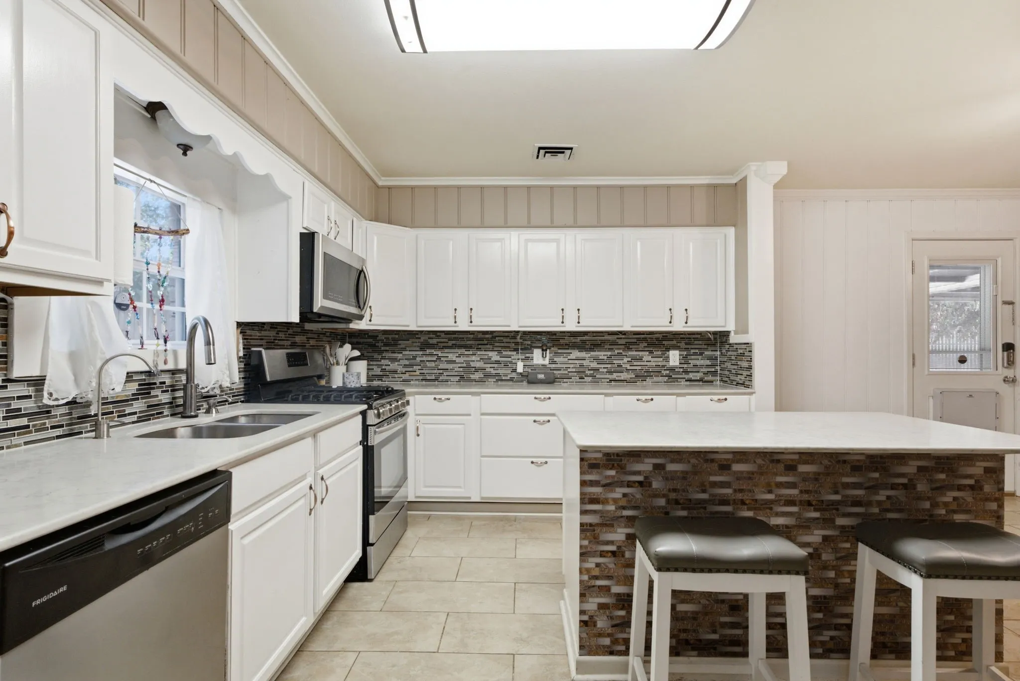White cabinetry with contrasting hardware, stainless steel appliances, mosaic tile backsplash, spacious island with stone-finish countertop, and tile flooring