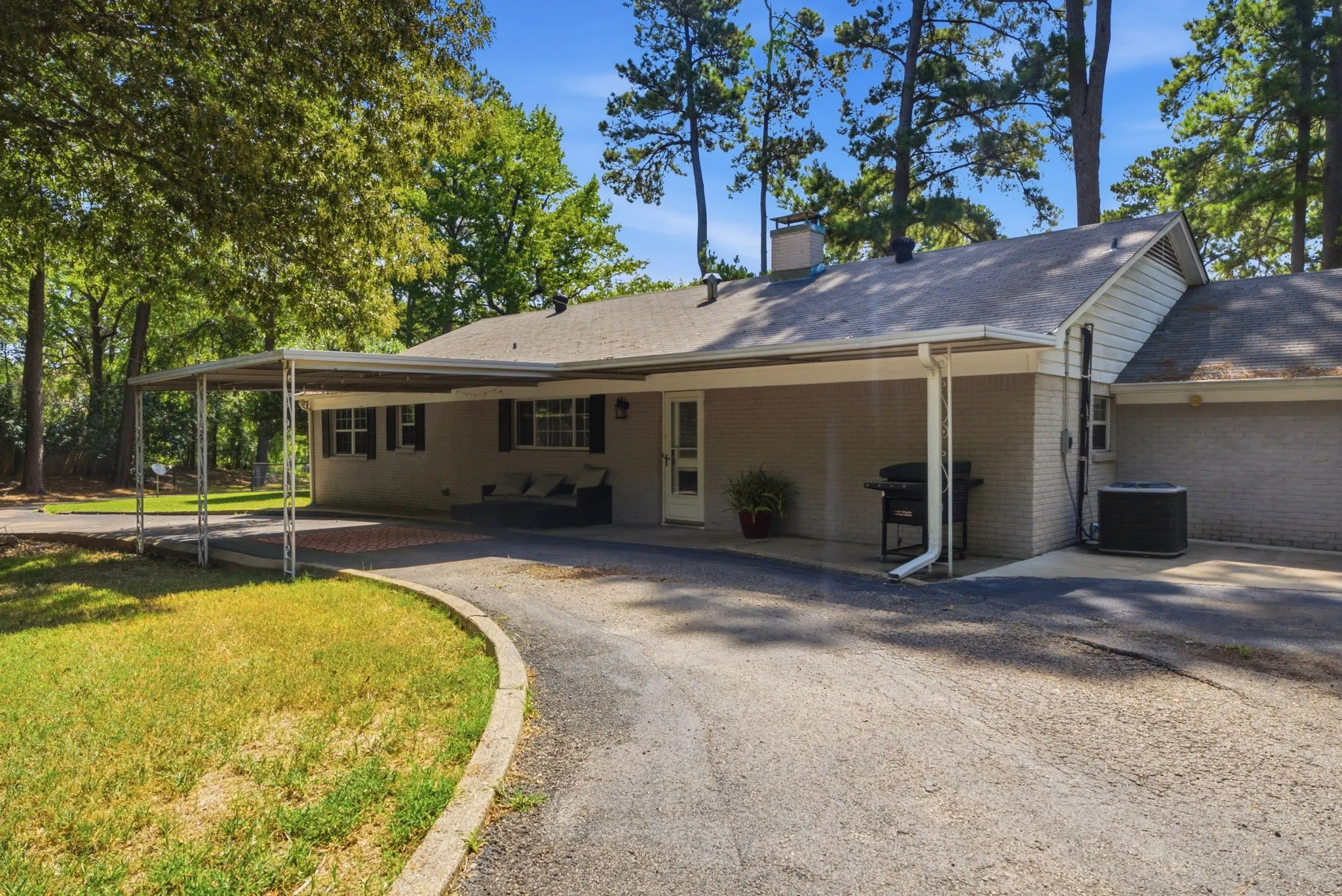 Gray brick exterior with a covered carport, asphalt driveway, and mature trees