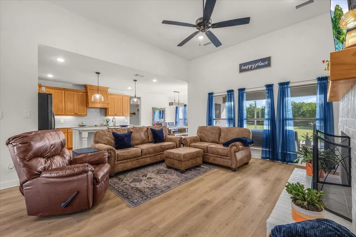 Living Room: Vaulted ceilings, a ceiling fan, abundant windows, and wood-look flooring complement the open layout connecting the living room and kitchen.