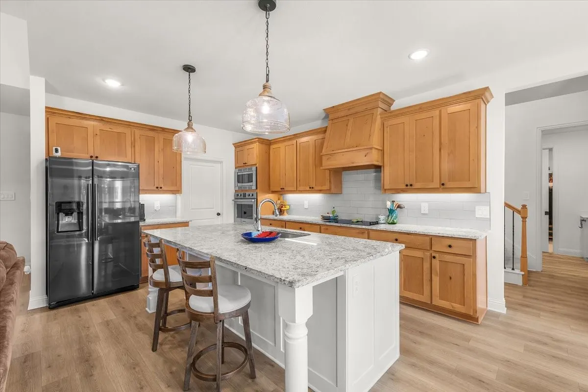 Kitchen: Spacious kitchen features warm wood cabinetry, light stone countertops, a contrasting island with seating, pendant lighting, and stainless appliances.