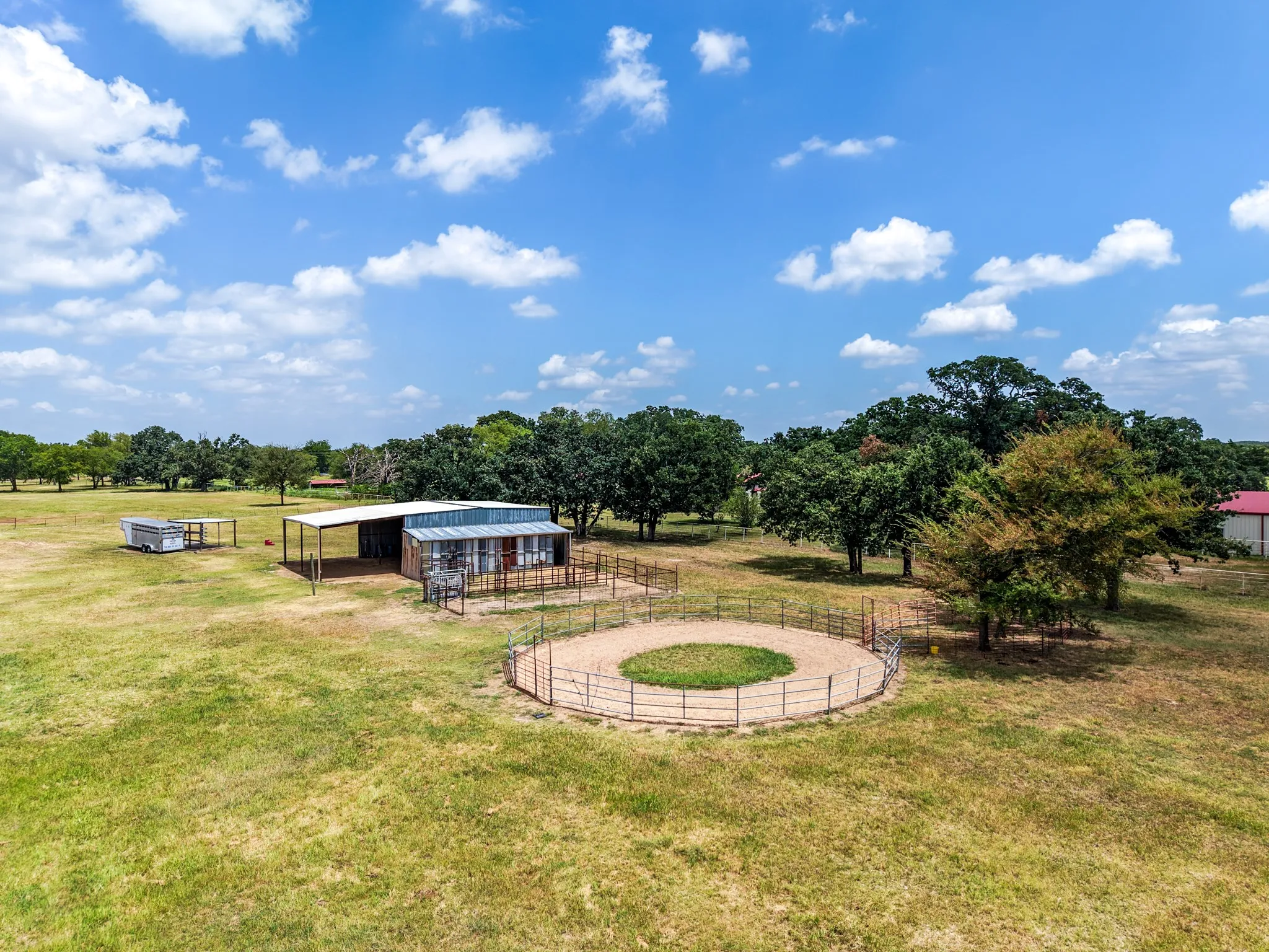 Expansive green space featuring a circular fenced arena, a barn with an attached open-sided shelter, and scattered mature trees