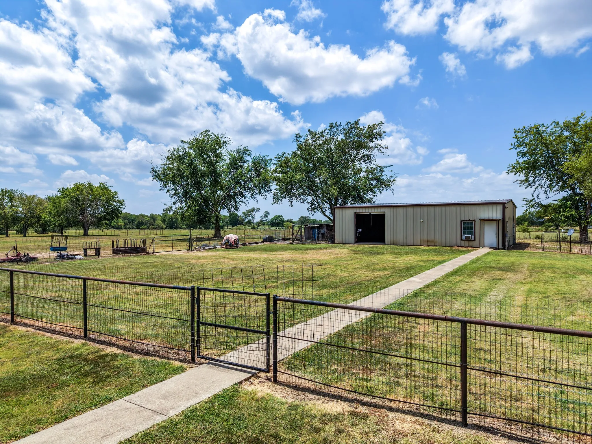 Expansive green space featuring a metal outbuilding with a roll-up door, a paved pathway, and a perimeter fence with a gate