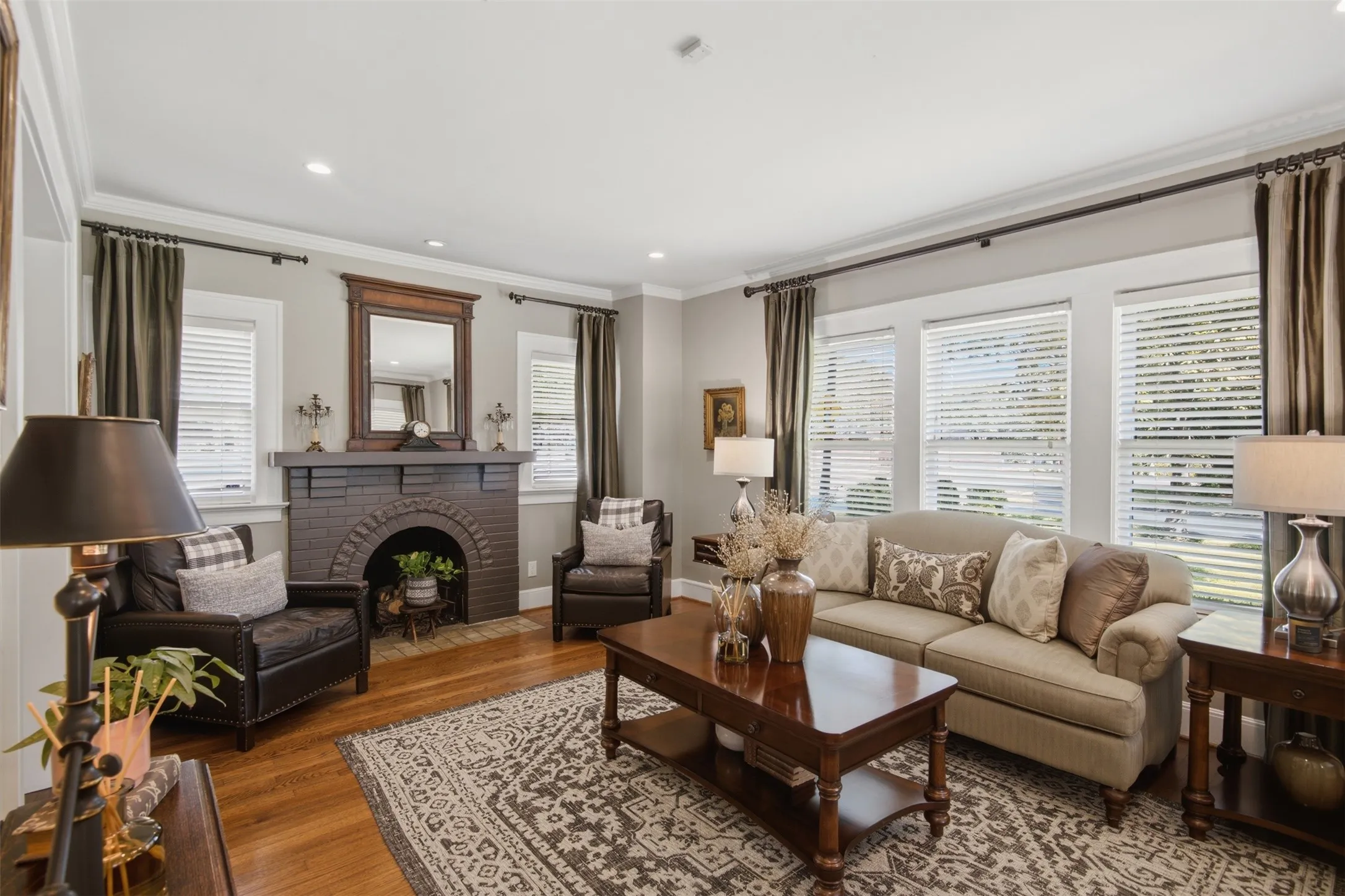 Warm living space featuring wood-finish flooring, a brick fireplace with an arched opening, multiple windows with blinds, recessed lighting, and crown molding