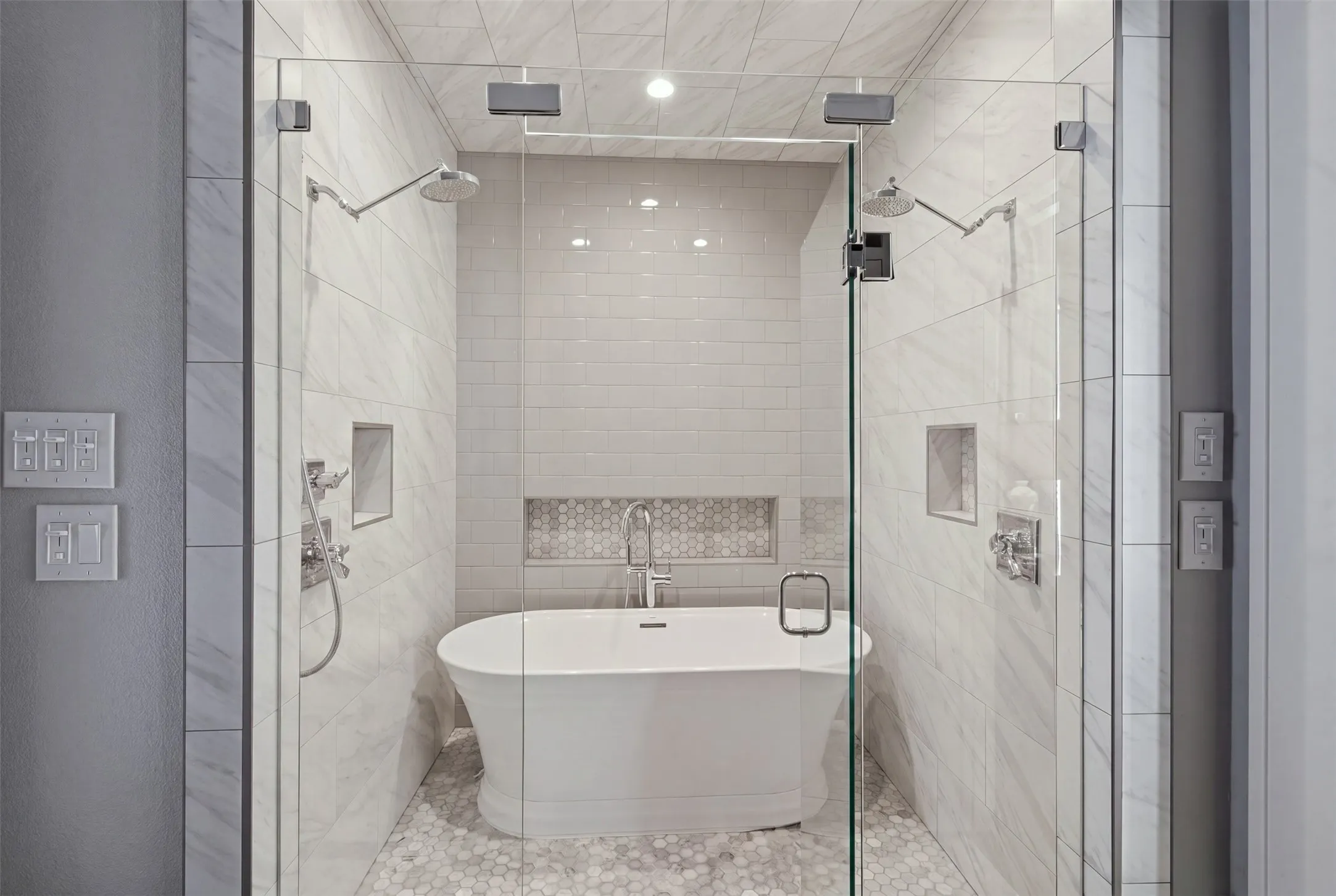 Glass-enclosed wet room featuring a freestanding soaking tub, dual rainfall showerheads, and integrated wall niches