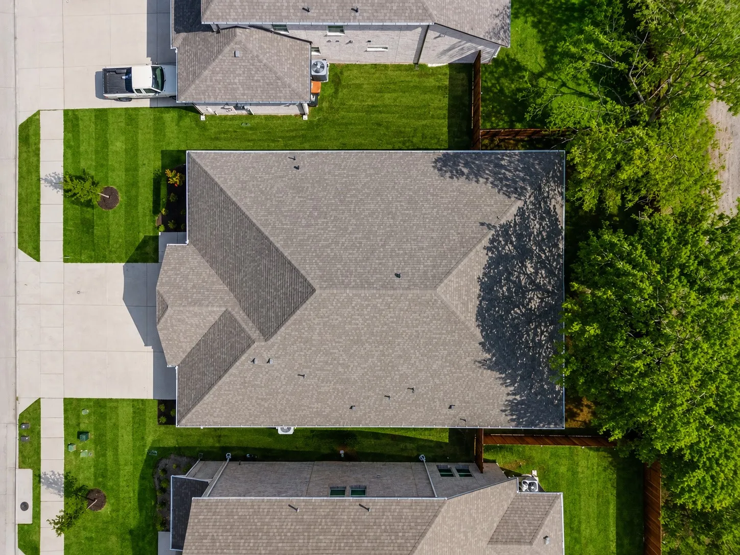Pitched roof with asphalt shingles, expansive green lawn, concrete pathways, mature trees bordering the property, and a wooden fence