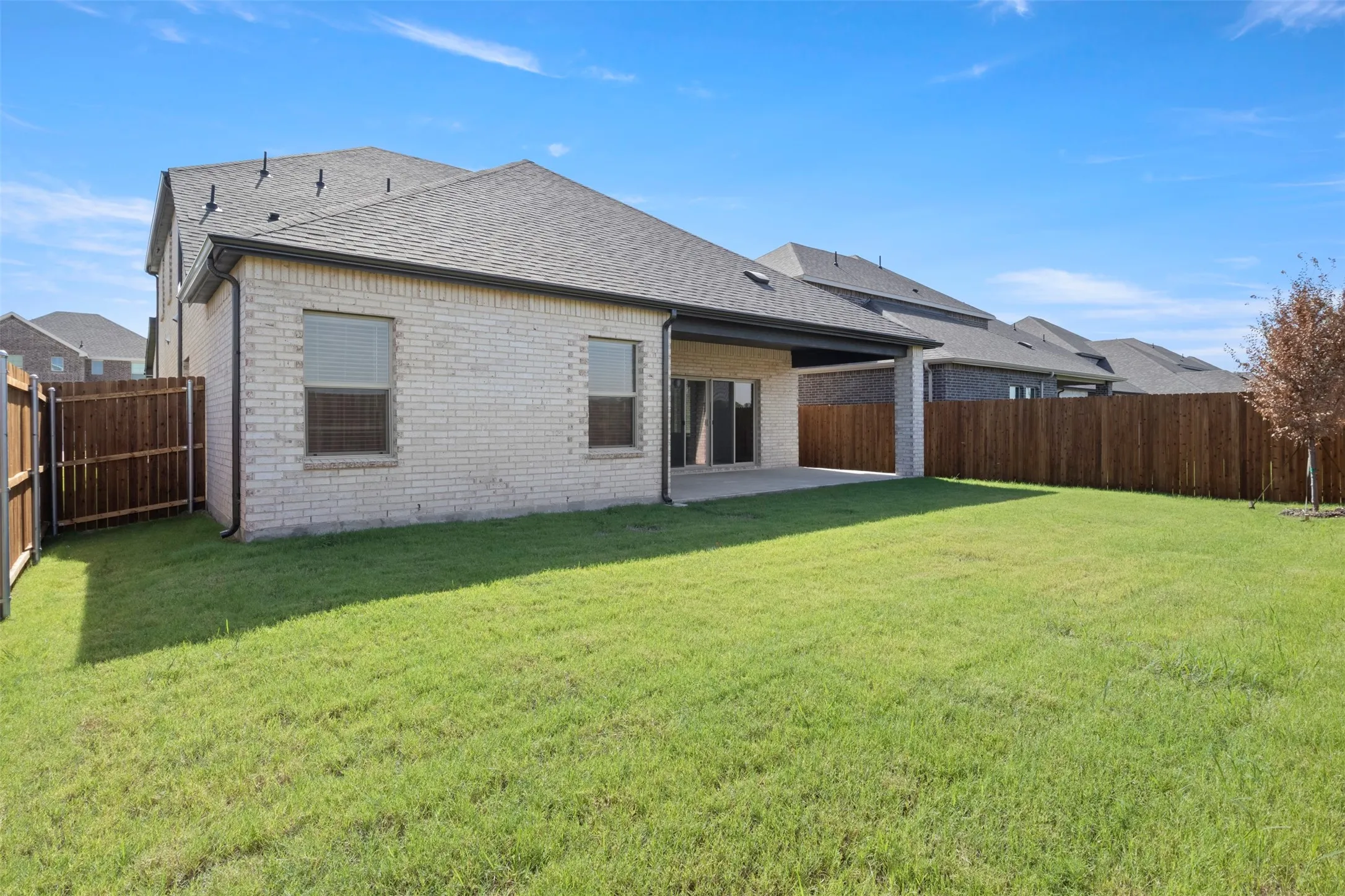 Expansive backyard featuring a covered patio, manicured lawn, full privacy fencing, and a light-toned brick exterior