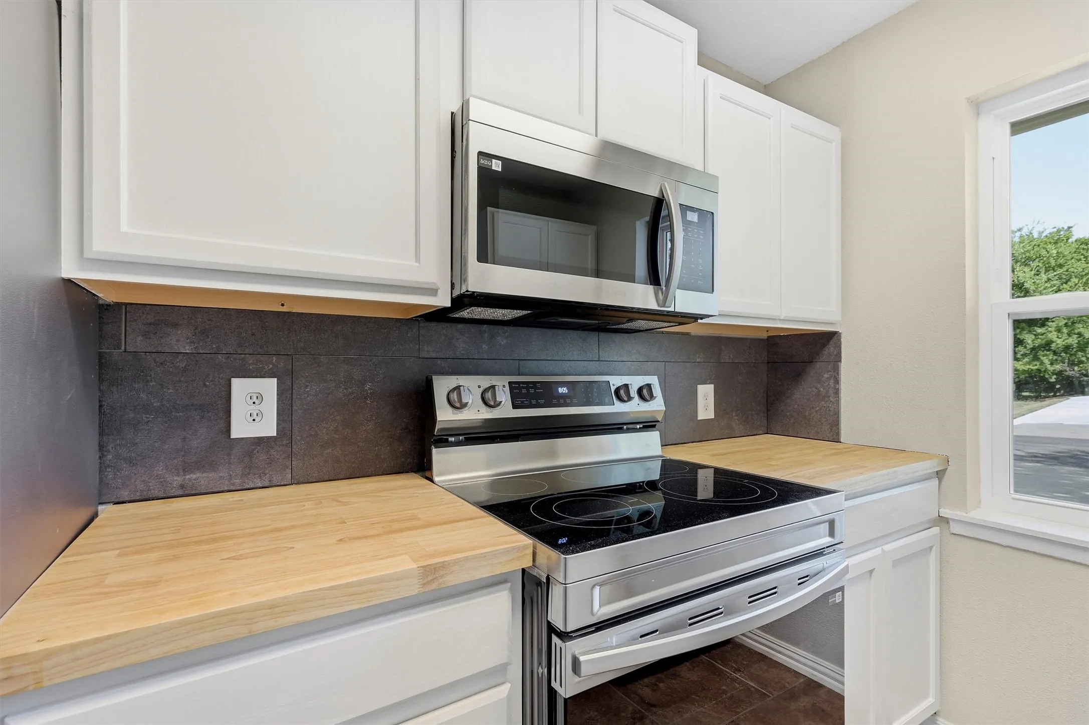White upper and lower cabinetry, stainless steel microwave, stainless steel range, light wood-finish countertops, and a dark tile backsplash