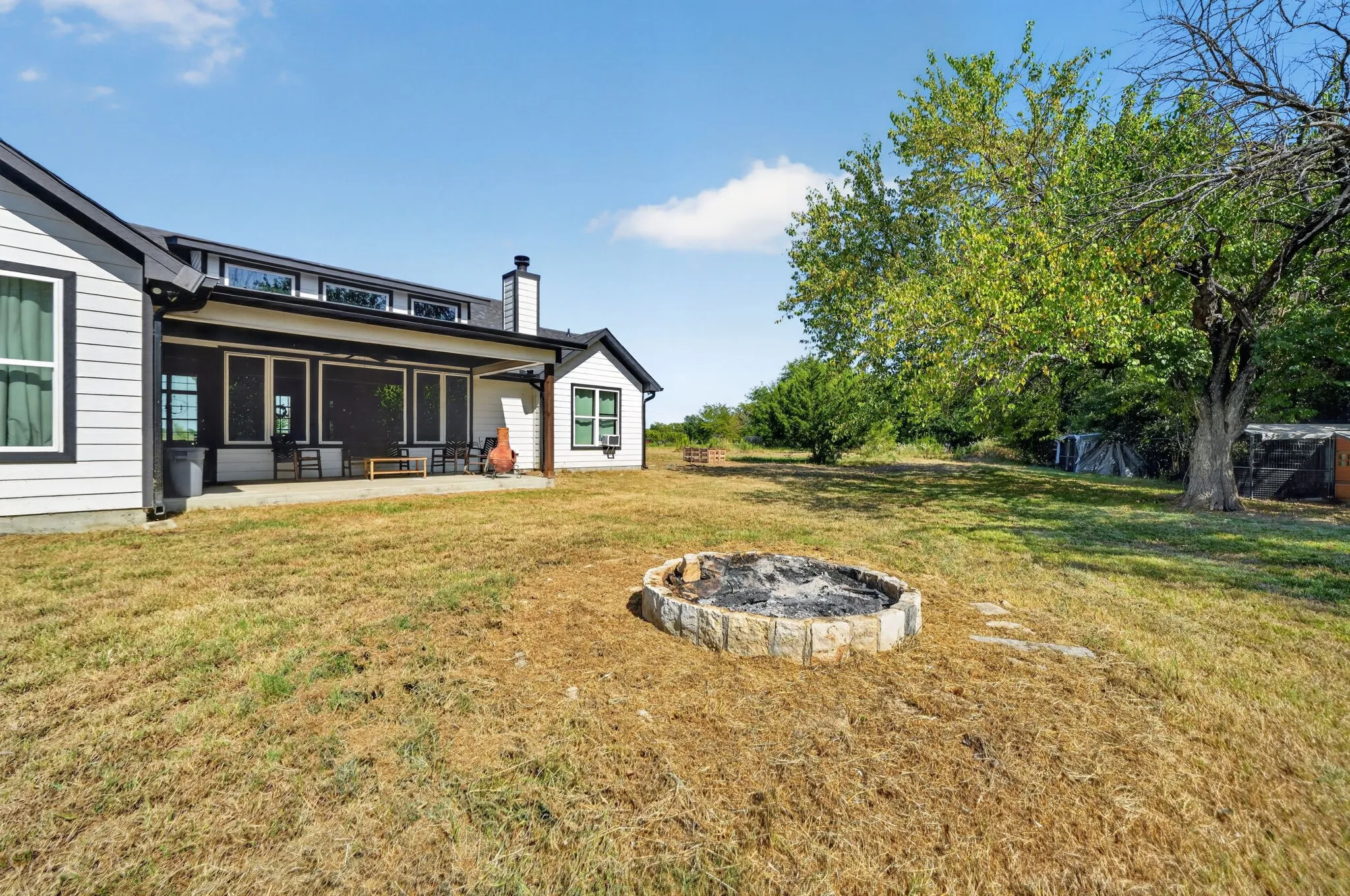 White siding exterior with black trim, covered patio, stone fire pit, and mature trees