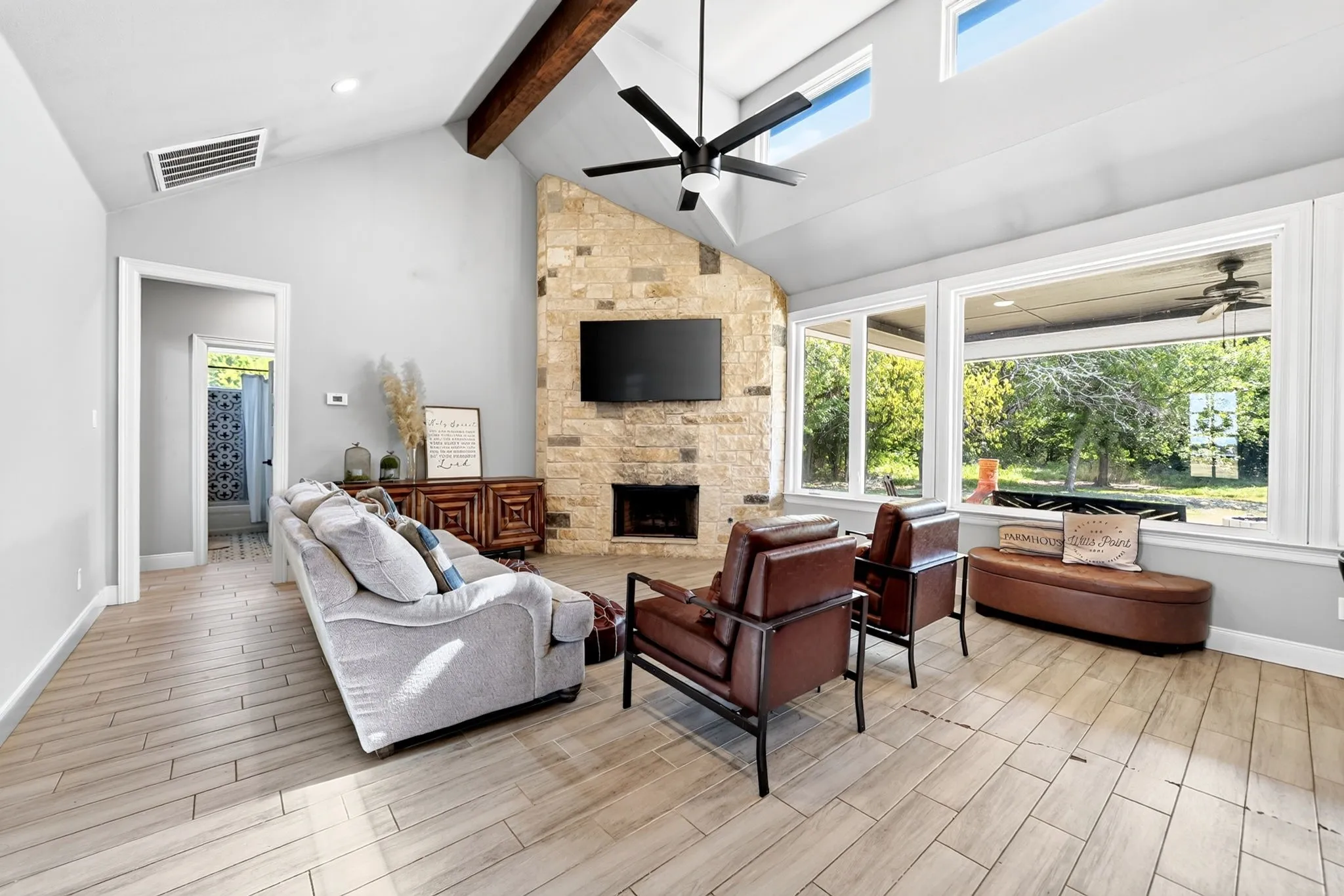 Vaulted ceiling with exposed wood beam, stone-clad fireplace, wood-finish flooring, large windows, and transom windows
