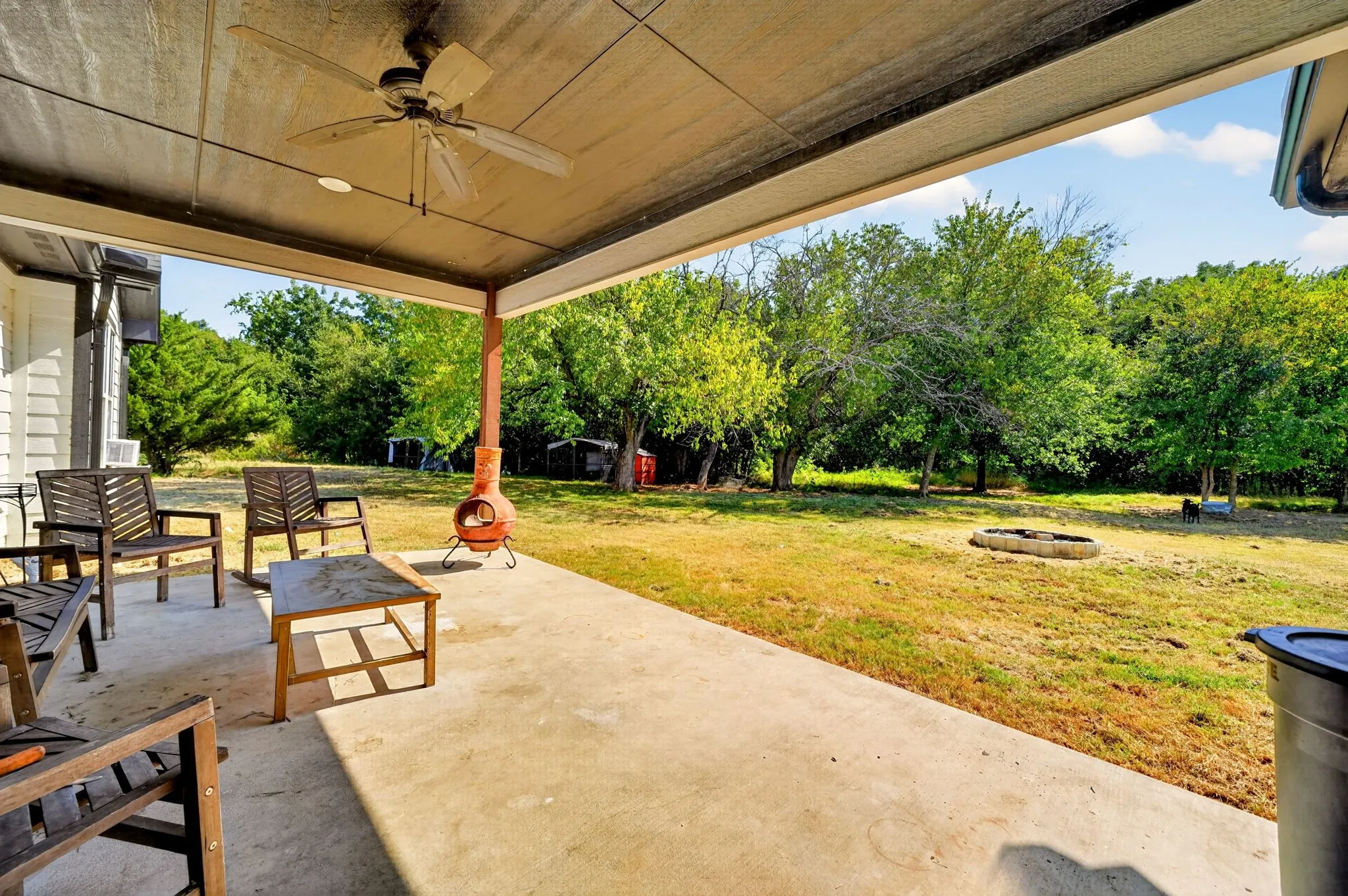Covered concrete patio with a ceiling fan, exposed wood support beams, and a spacious grassy yard beyond