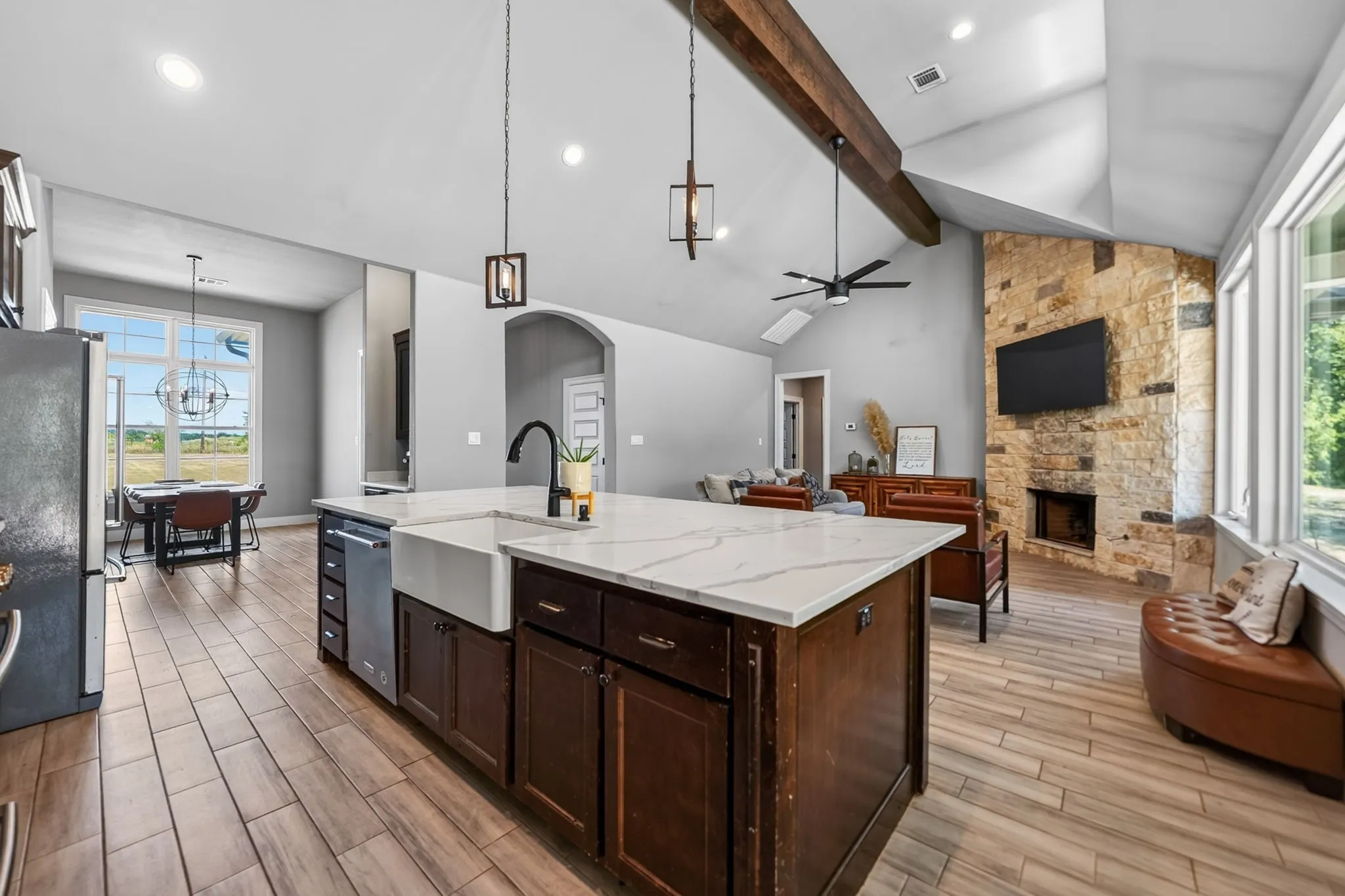 Vaulted ceilings with exposed wood beams, a stone fireplace, wood-finish flooring, a kitchen island with a farmhouse sink, and a multi-pane window