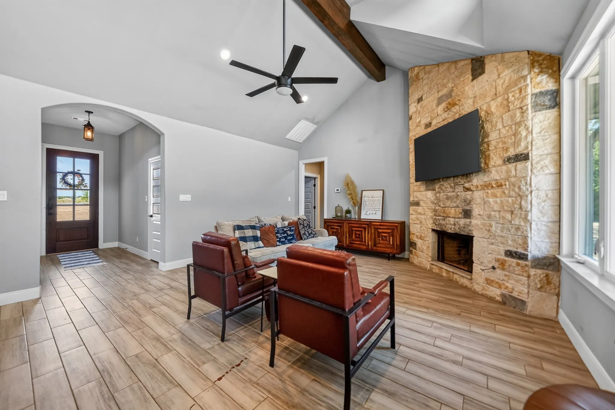 Vaulted ceiling with exposed wood beam, corner stone-finish fireplace, wood-finish flooring, ceiling fan, and an arched entry