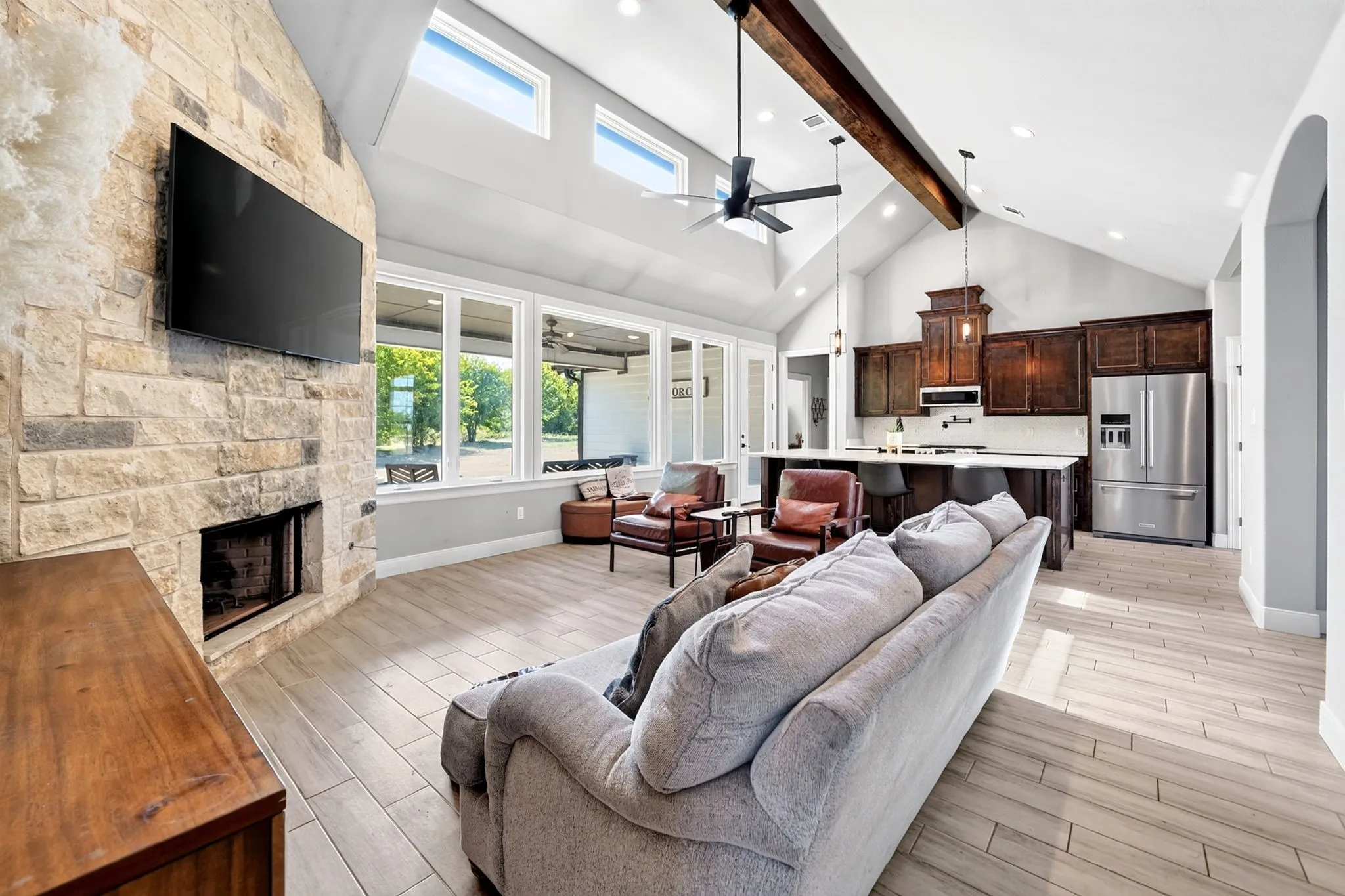 Vaulted ceiling with exposed wood beam, a stone-clad fireplace, large windows, wood-finish flooring, and a kitchen featuring dark wood cabinetry and stainless steel appliances