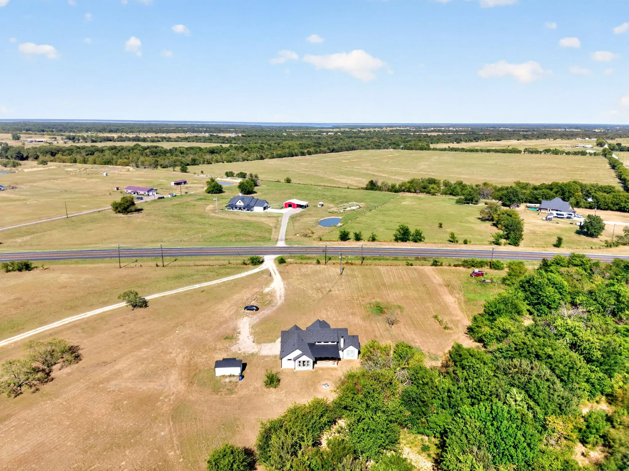 Expansive property featuring a modern home with a dark roof and white exterior, mature trees, open fields, and a paved road