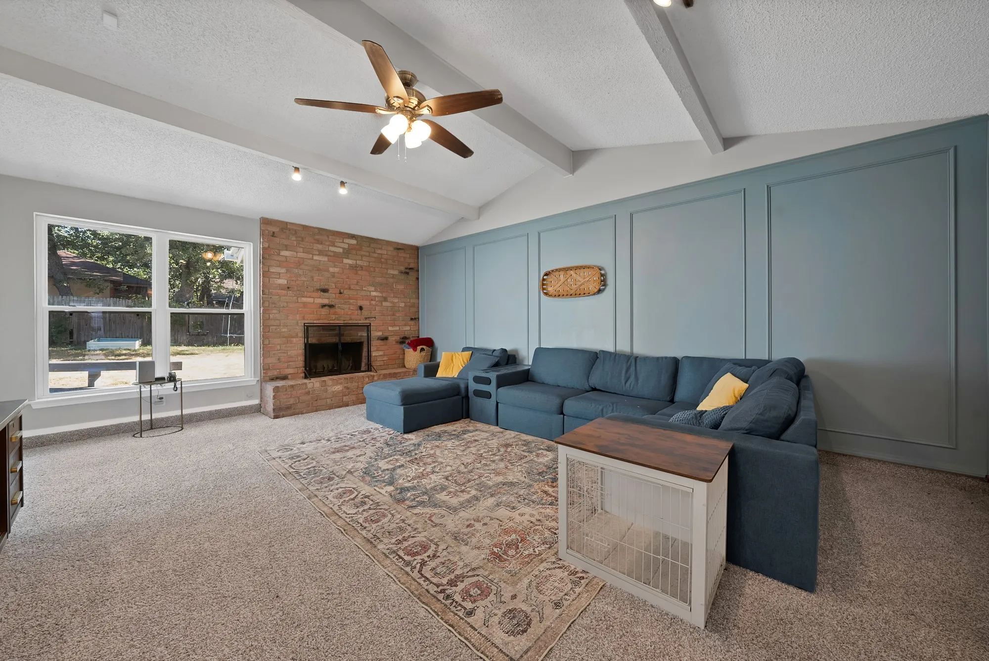 Vaulted ceiling with exposed beams, brick fireplace, large picture windows, wood-finish ceiling fan, and panel accent wall