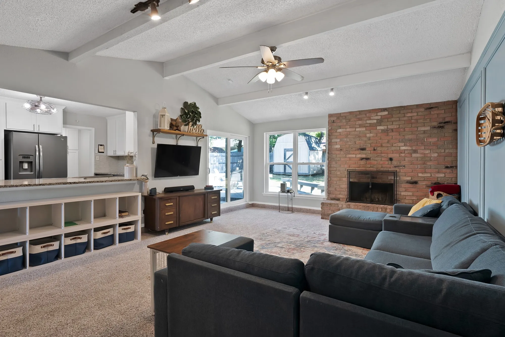 Vaulted ceiling with exposed beams, brick fireplace, open-concept kitchen pass-through, built-in shelving, and direct outdoor access
