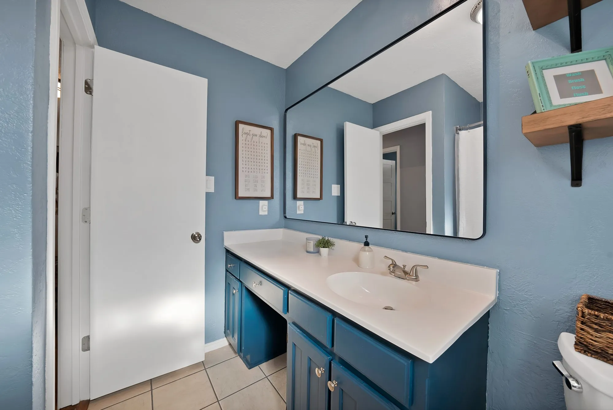 Vanity with integrated sink, expansive mirror, wood-finish floating shelf, and neutral tile flooring