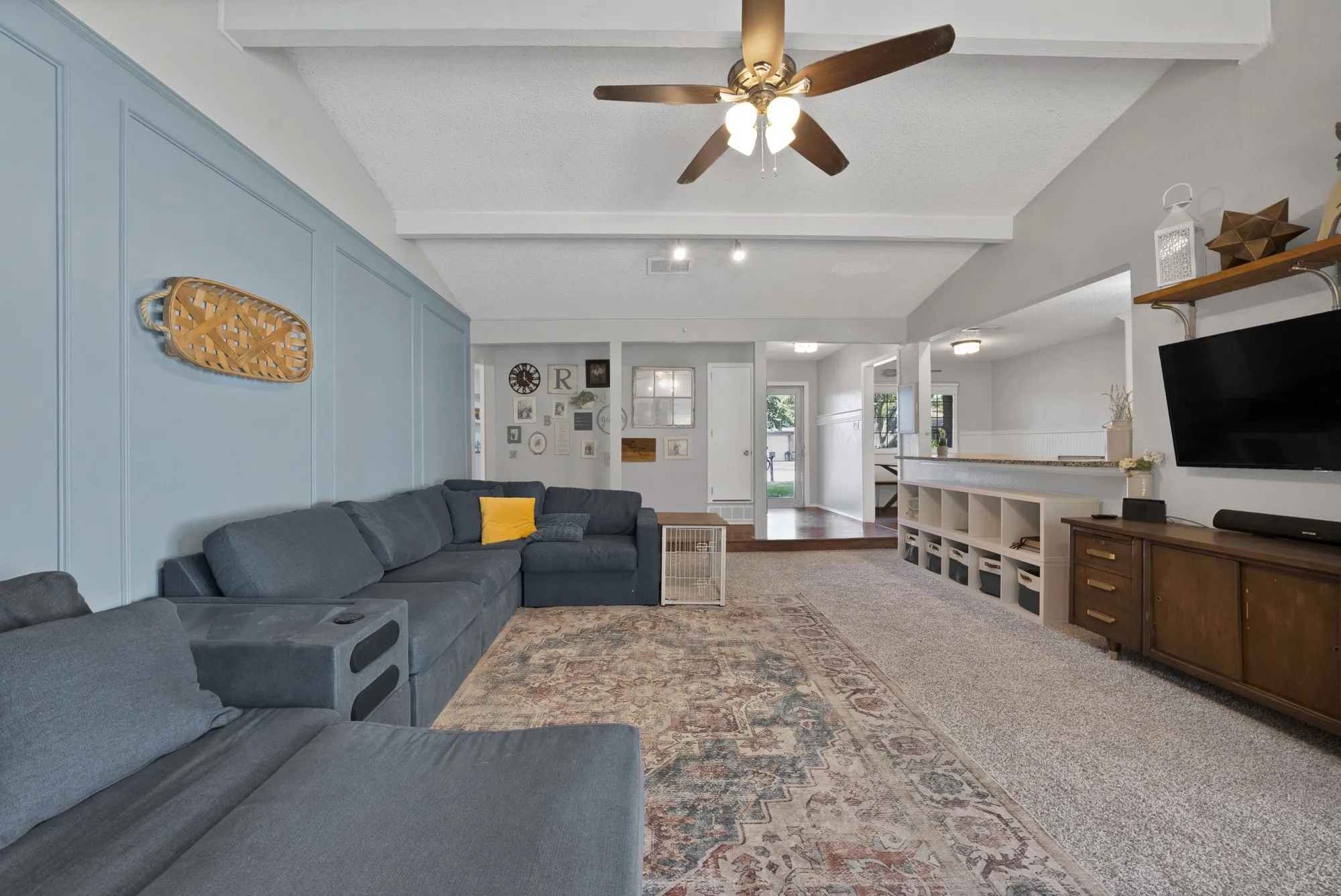 Vaulted ceiling with exposed beams, wood-finish ceiling fan, accent wall with panel molding, built-in shelving, and recessed lighting