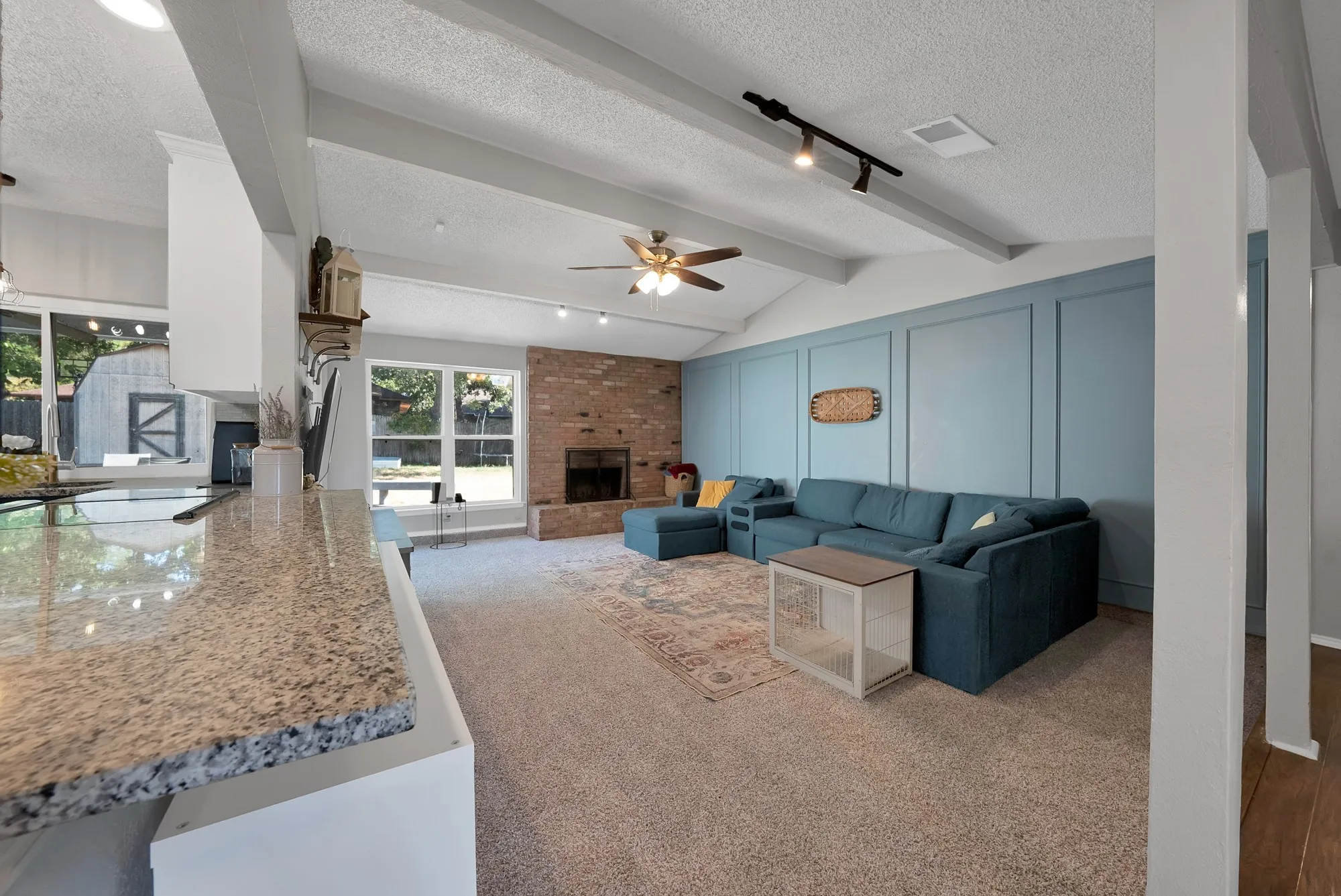 Vaulted ceiling with exposed beams, brick fireplace, expansive window, accent wall paneling, and granite countertop