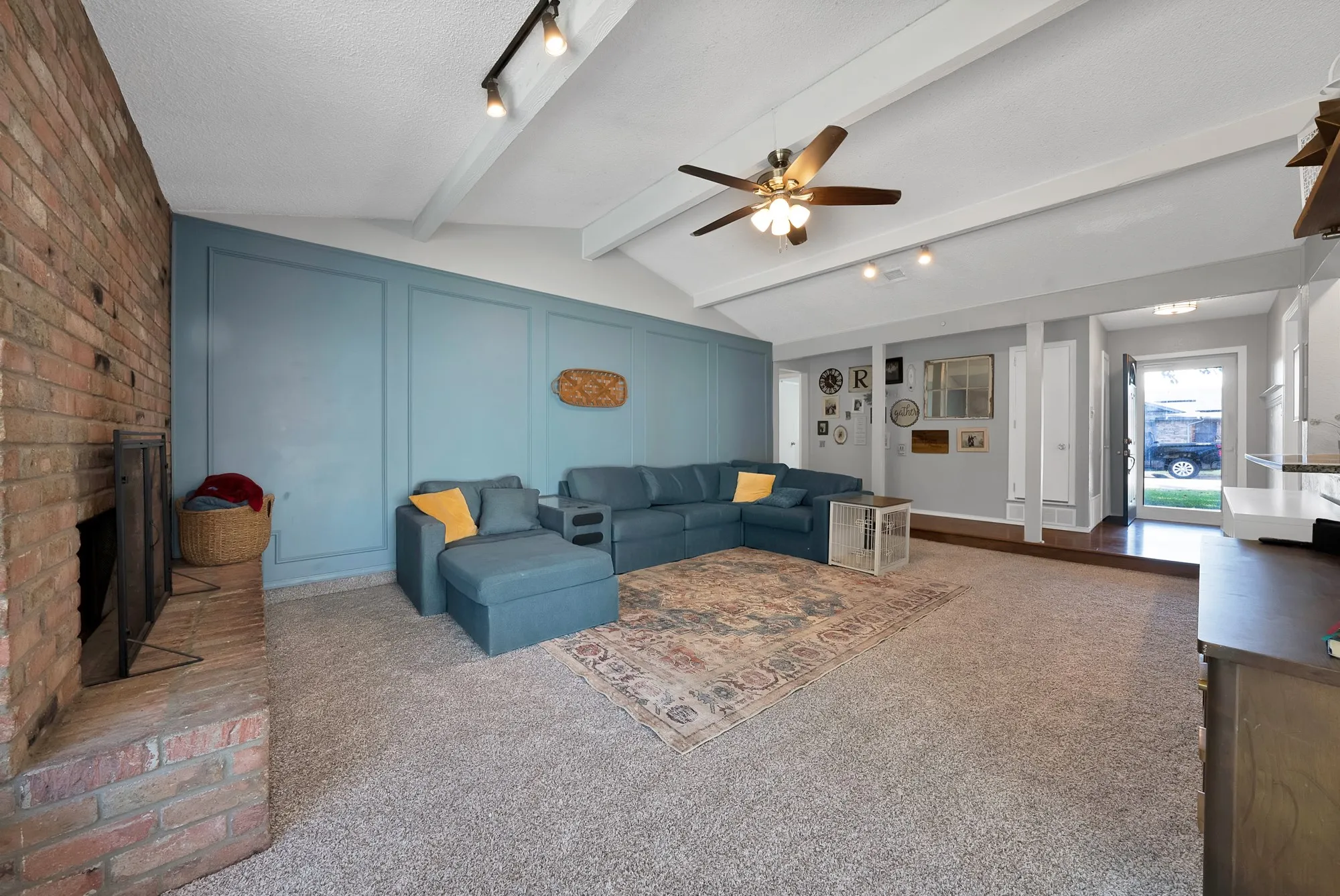 Vaulted ceiling with exposed beams, brick fireplace, accent wall with panel molding, ceiling fan, and track lighting