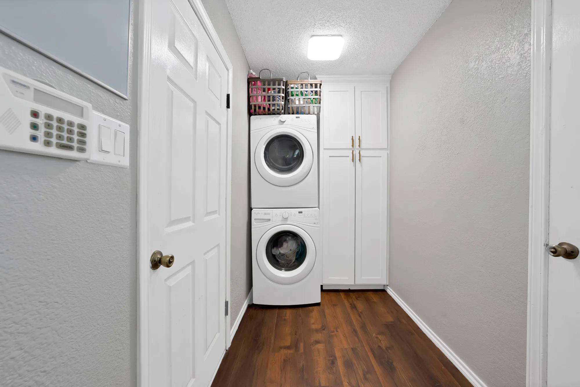 Stacked washer and dryer, wood-finish flooring, white cabinetry with gold-tone hardware, textured walls, and a ceiling-mounted light fixture
