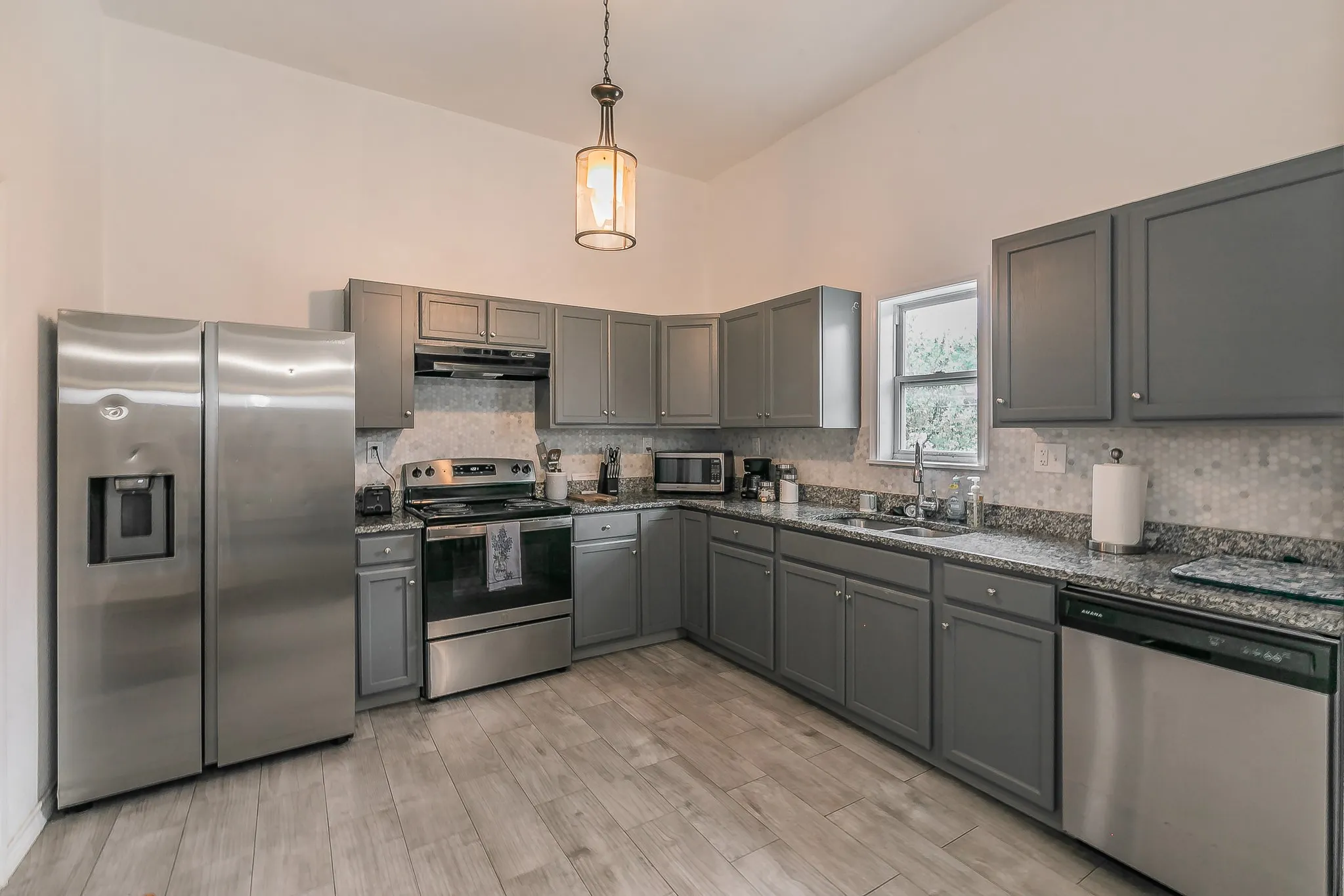 Kitchen featuring wood-finish flooring, granite countertops, gray cabinetry, stainless steel appliances, and a mosaic tile backsplash
