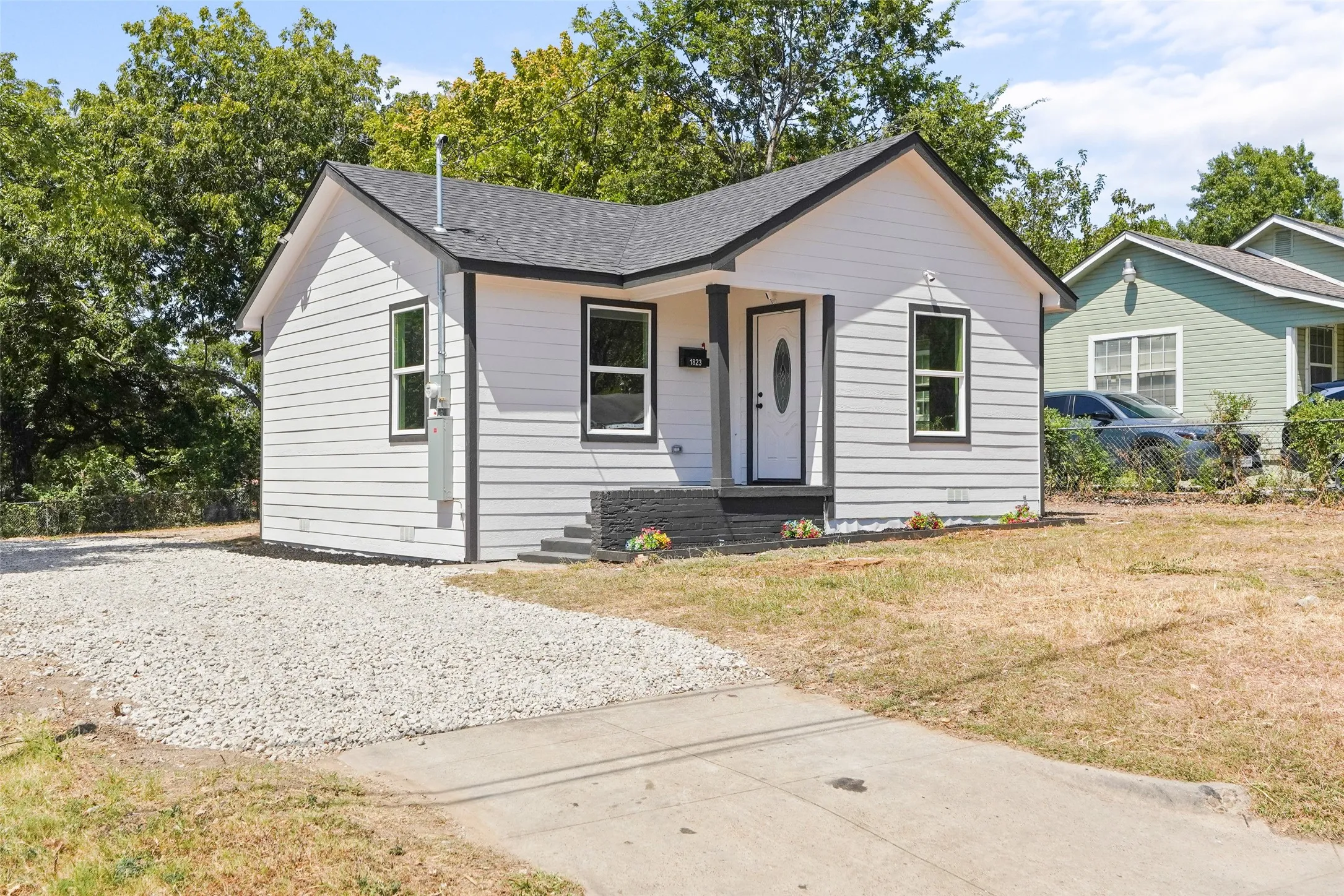 White siding exterior with dark trim accents, black shingle roof, white entry door with oval glass insert, and a gravel driveway