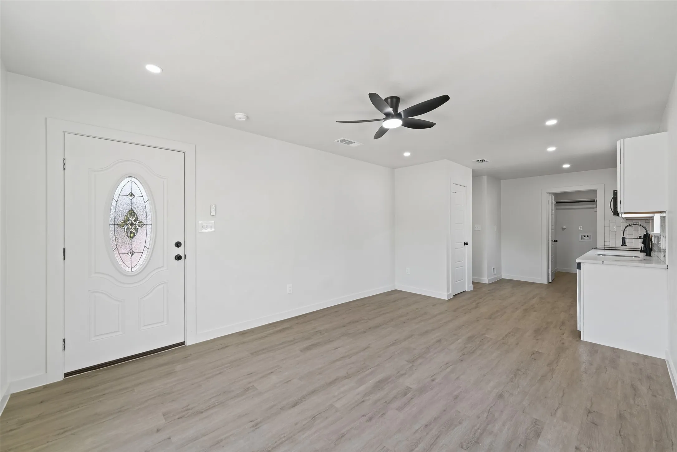 Wood-finish flooring, recessed lighting, matte black ceiling fan, white paneled door with leaded glass insert, and a compact kitchen area with white cabinetry