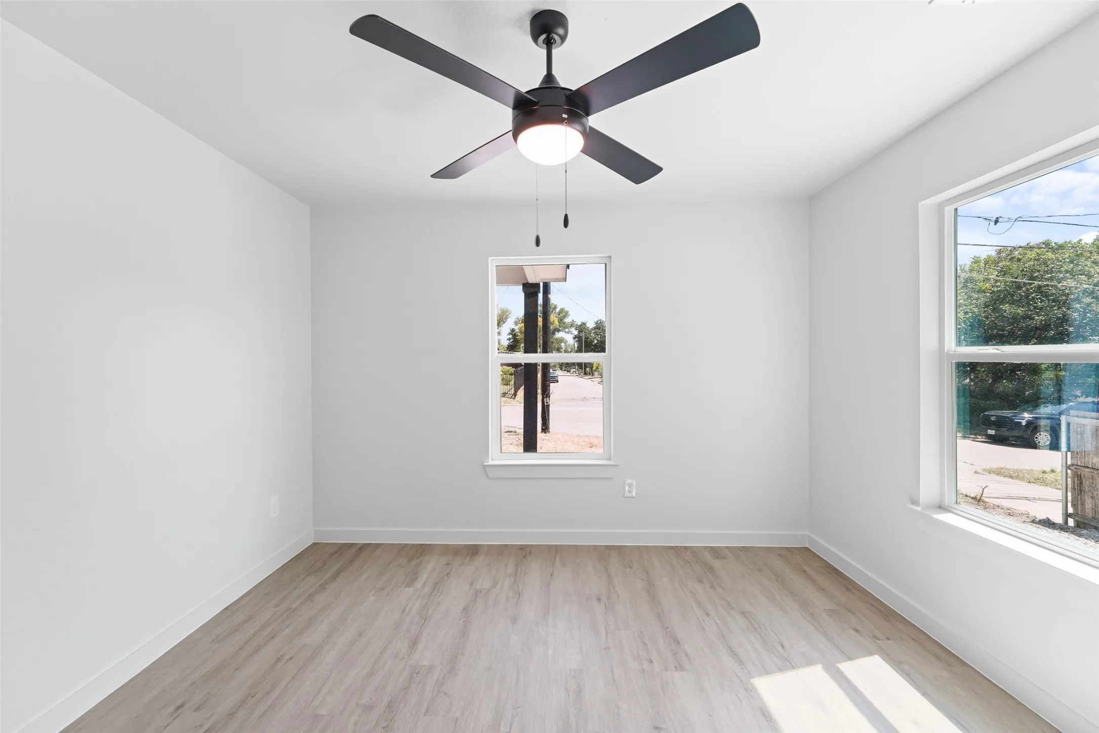 Light wood-finish flooring, two large windows, a ceiling fan with integrated lighting, and crisp white walls